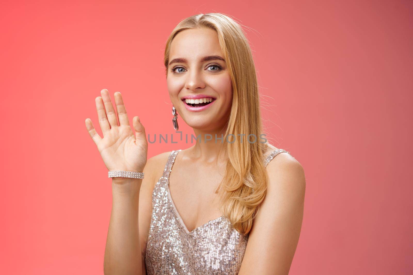 Waist-up studio shot friendly attractive elegant tender blond woman say hi waving raised hand greeting welcoming friend smiling delighted introducing herself hello gesture, red background.