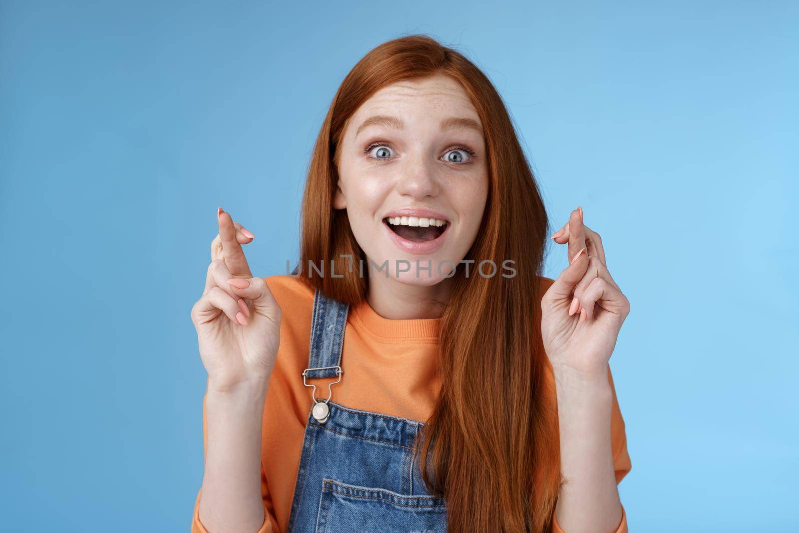 Girl realising prayers fulfilled dreams come true hearing good news standing surprised amused smiling broadly wide eyes impressed cross fingers good luck finally desire wish achieved, blue background.