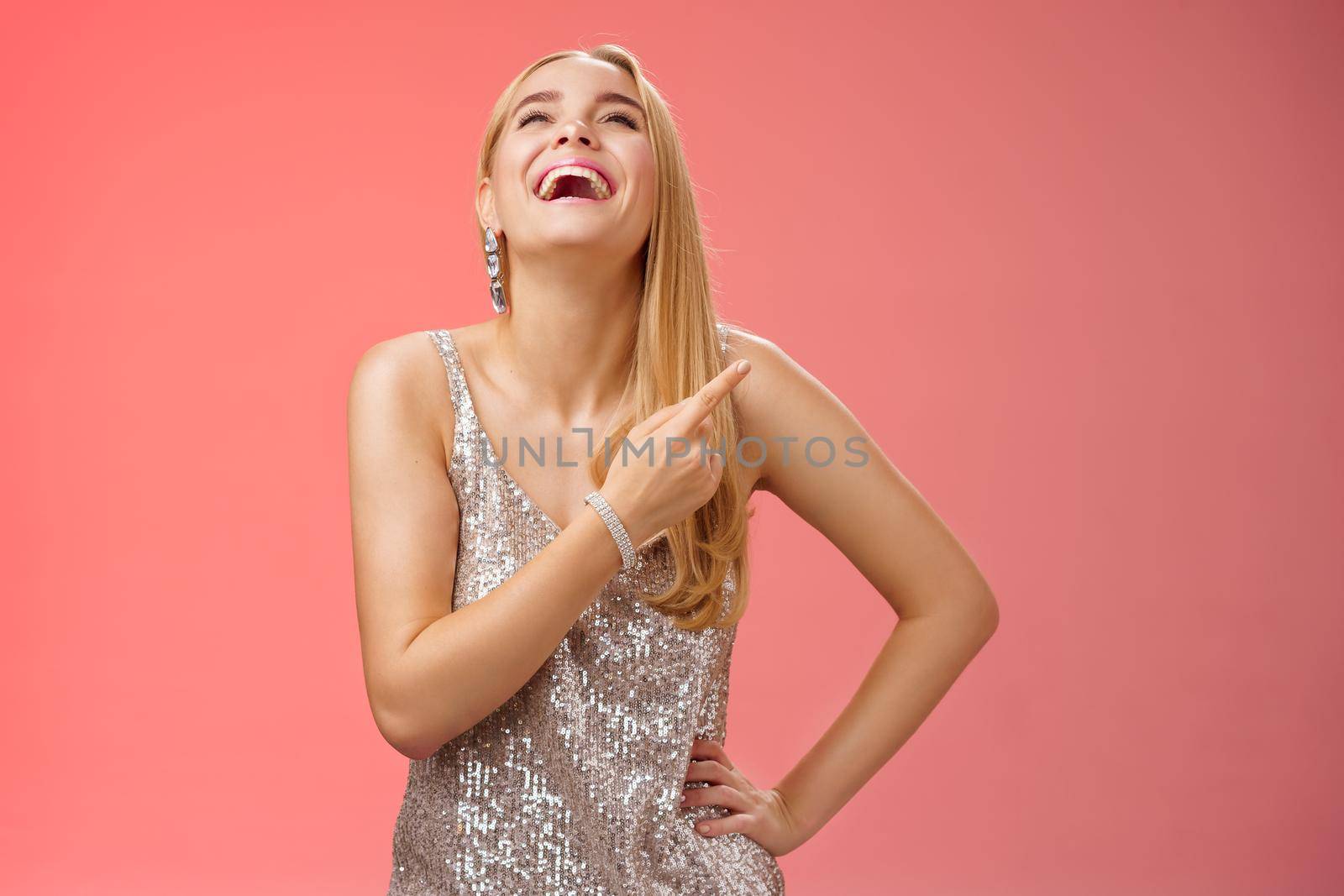 Fabulous carefree attractive blond woman in silver evening party dress laughing out loud have fun raise hand joyfully up pointing right index finger enjoying awesome sense humour, red background.