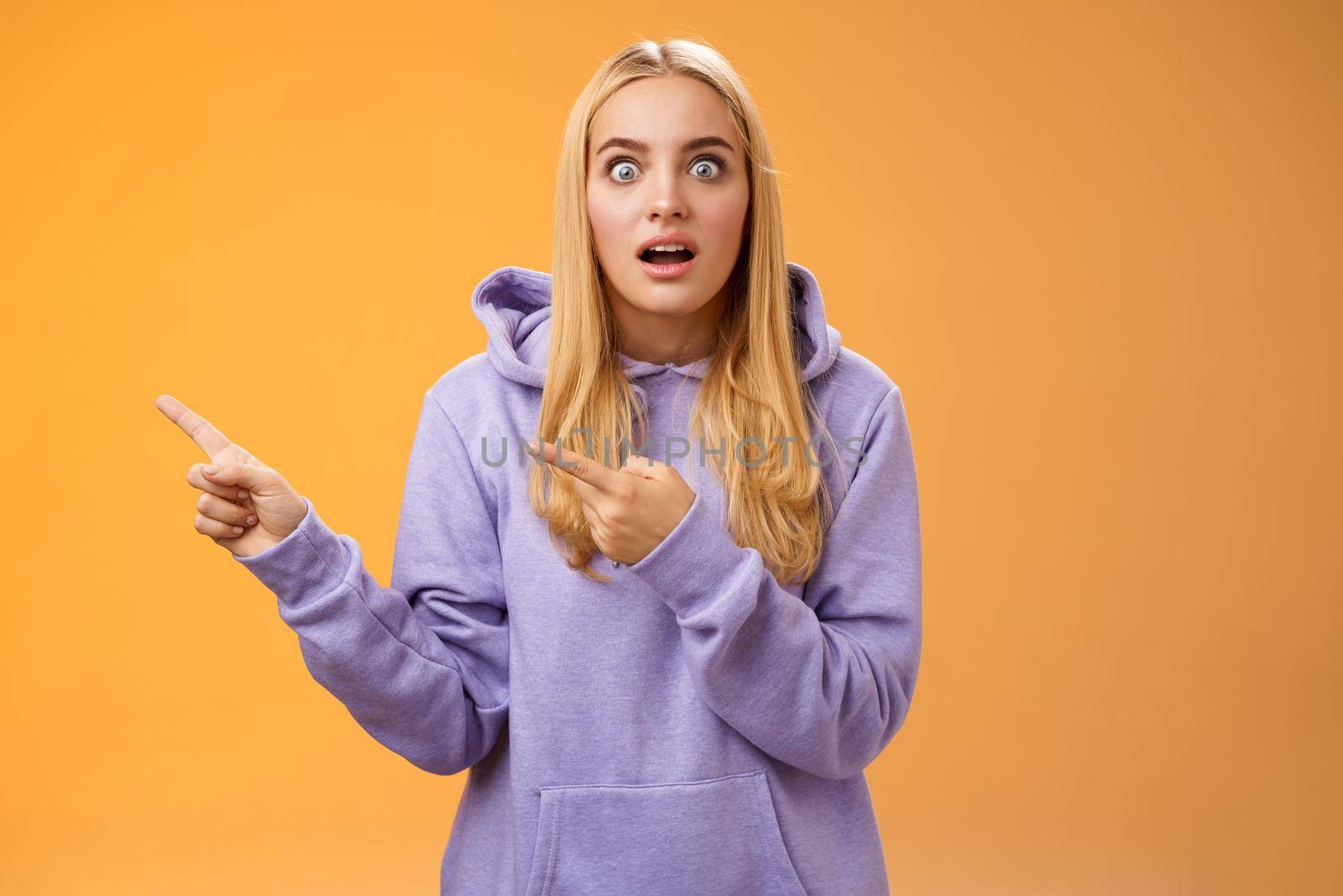 Wow astonished impressed good-looking blond european female in hoodie pop eyes wondered surprised questioned drop jaw pointing left excited asking if gossip true, standing orange background by Benzoix