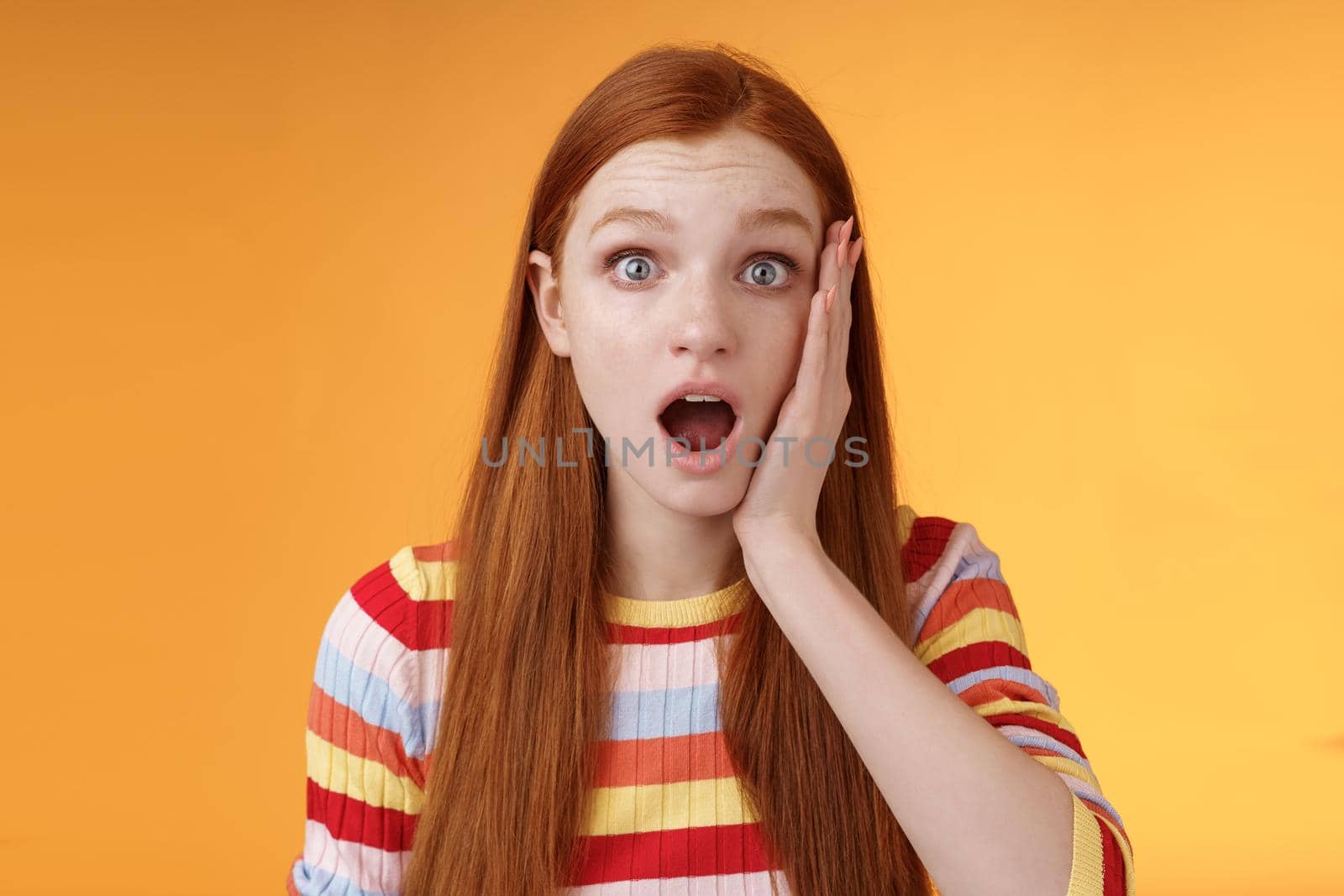 Shocked impressed concerned ginger girl drop jaw gasping stunned slap cheek wide eyes surprised hear disturbing terrible news standing orange background sympathizing awful story by Benzoix