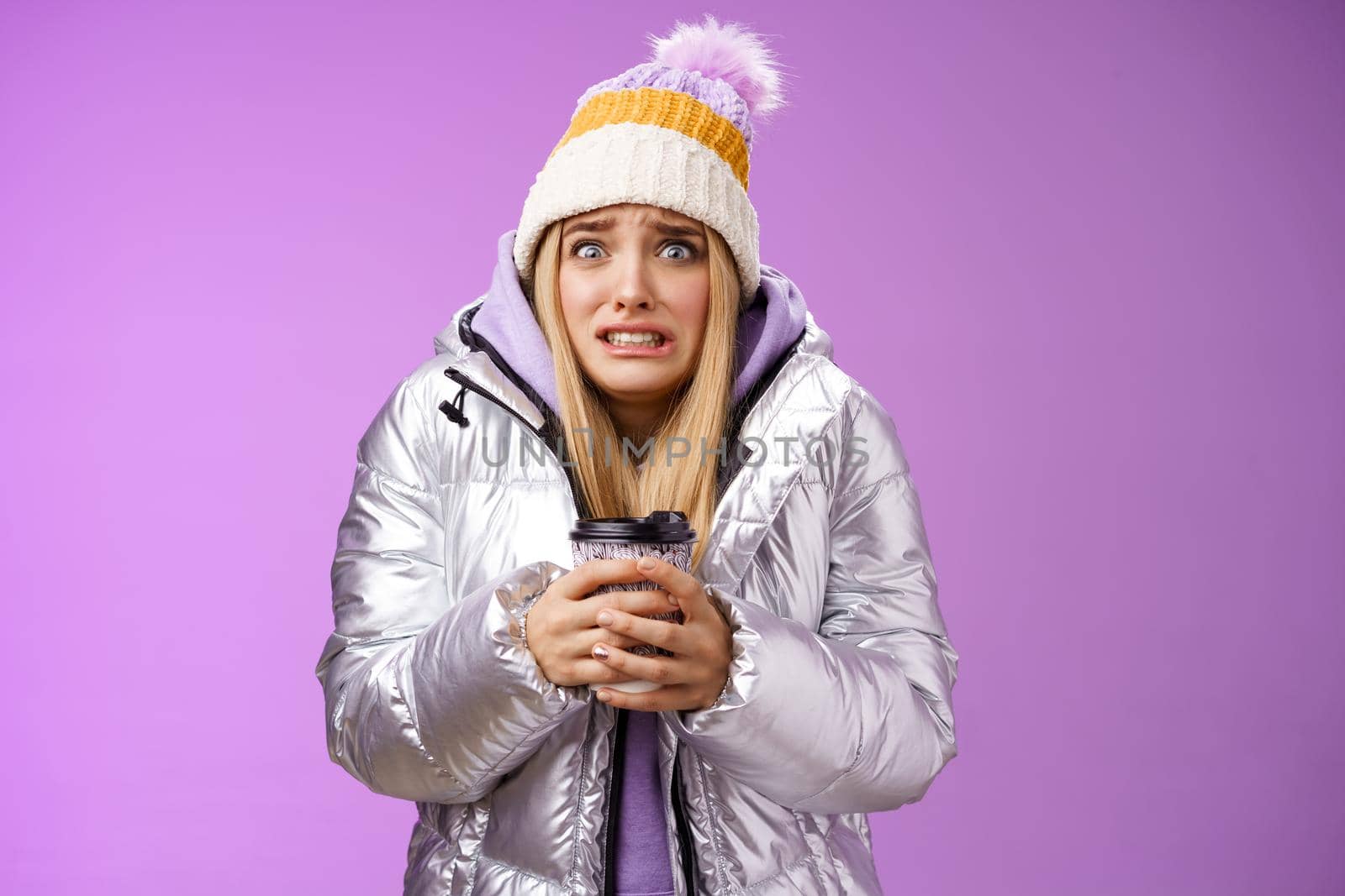 Shaking uncomfortable displeased girl wearing stylish silver jacket hat trembling freezing cold warm hands holding take-away coffee cup clench teeth pop eyes discomfort, standing purple background by Benzoix