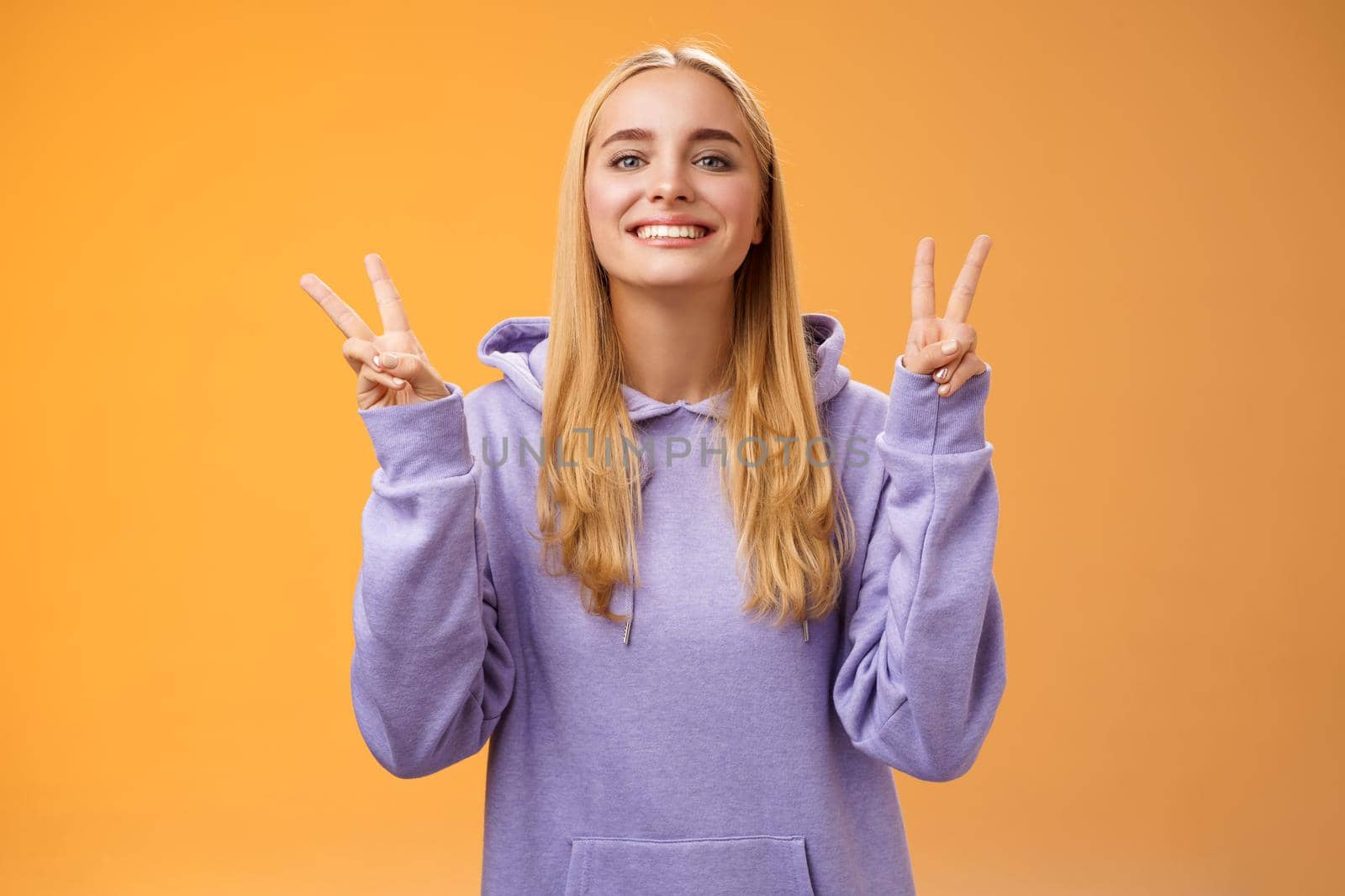 Lifestyle. Peaceful glad attractive blond european young female college student in hoodie smiling friendly make peace victory gesture spread love and happiness standing hippie sign orange background.