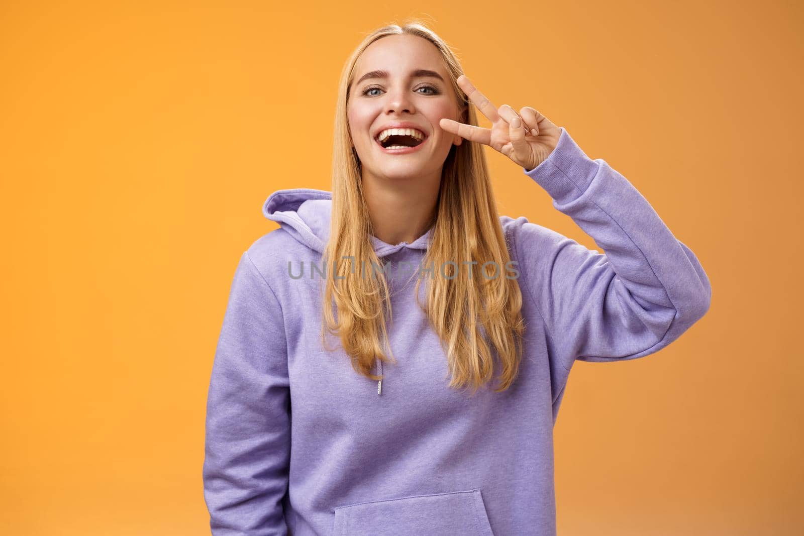 Attractive friendly carefree millennial blond girl in purple hoodie having fun friends laughing joyfully show peace victory hippie sign adore perfect chilly spring weather, orange background by Benzoix