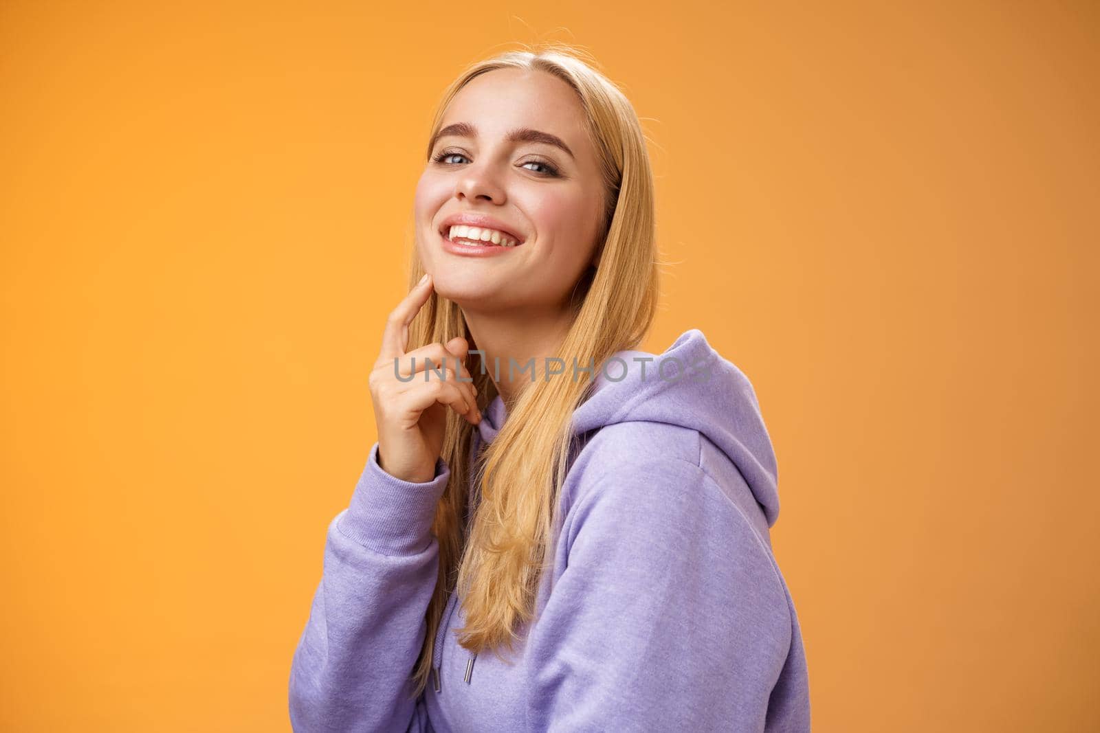 Attractive sassy self-assured caucasian blond female in purple hoodie turning camera touching chin cheeky smile camera check out interesting thing gazing forward intrigued, standing orane background by Benzoix