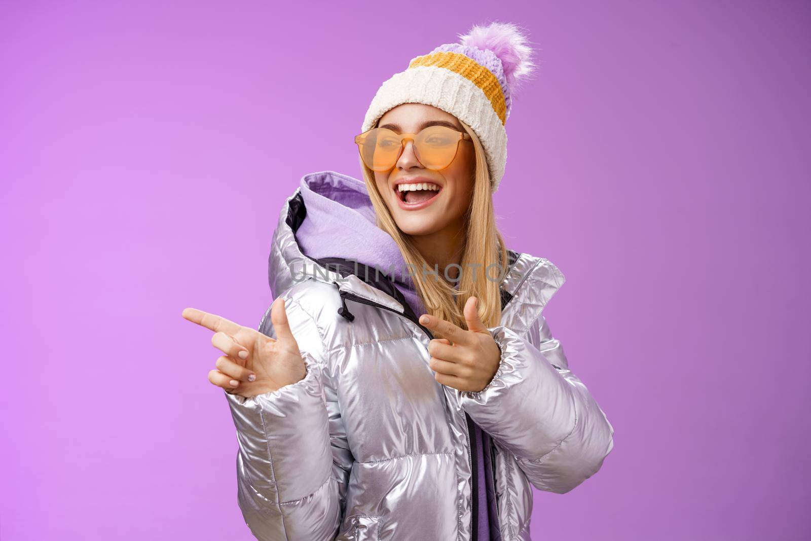 Hey how you doin. Cheeky stylish bond girl having fun greeting friend pointing finger pistols left smiling sassy say hello what up wearing cool silver jacket hat sunglasses, purple background.