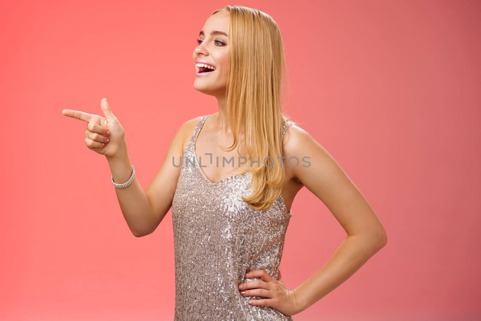 Stylish gorgeous young blond caucasian girlfriend in silver stylish evening dress turning profile left pointing curiously smiling broadly enjoying concert perfomance during party, red background.