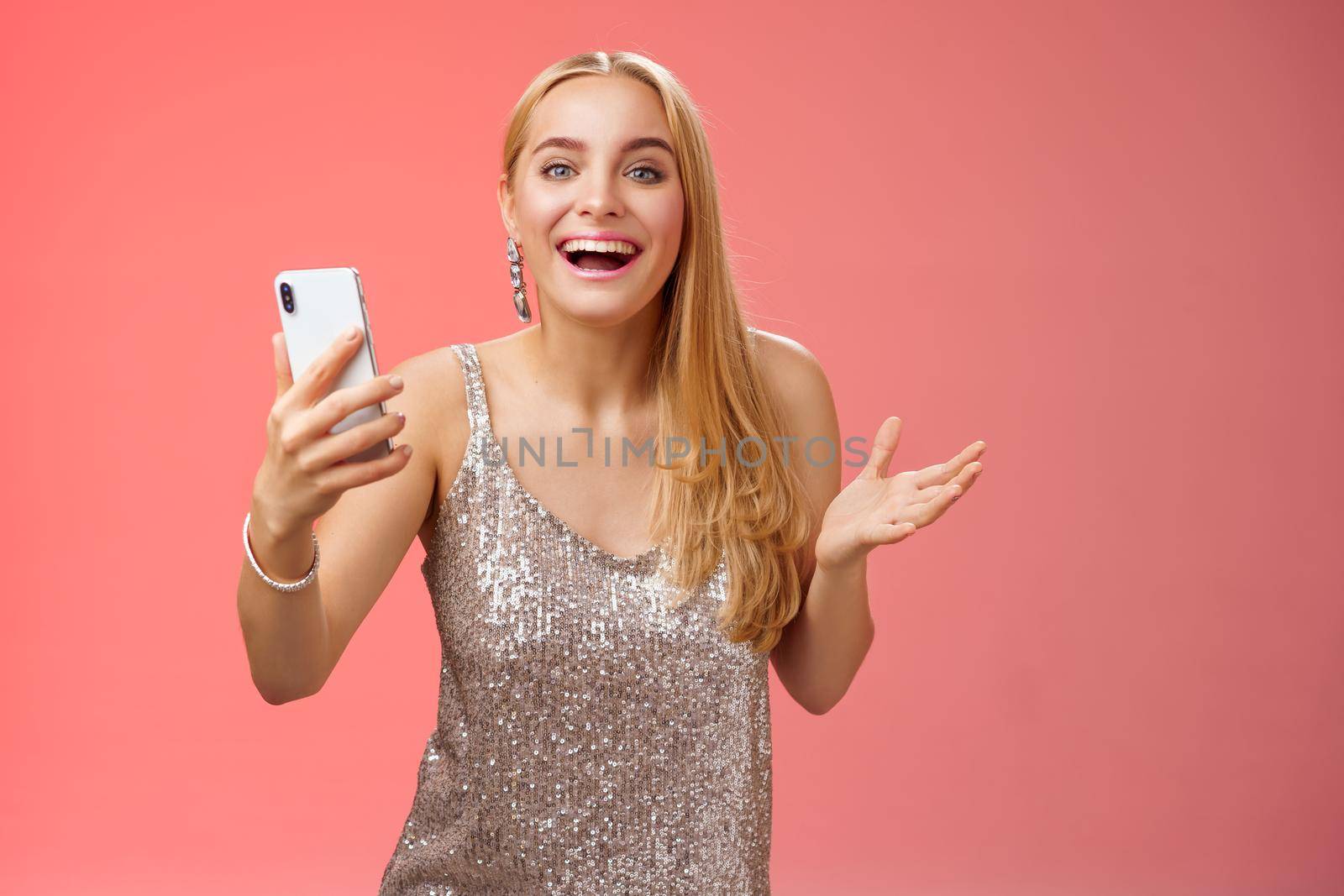 Astonished happy charming blond girl in silver glittering stylish dress holding smartphone amazed liking awesome result edit photo app smiling wondered amused, standing red background by Benzoix