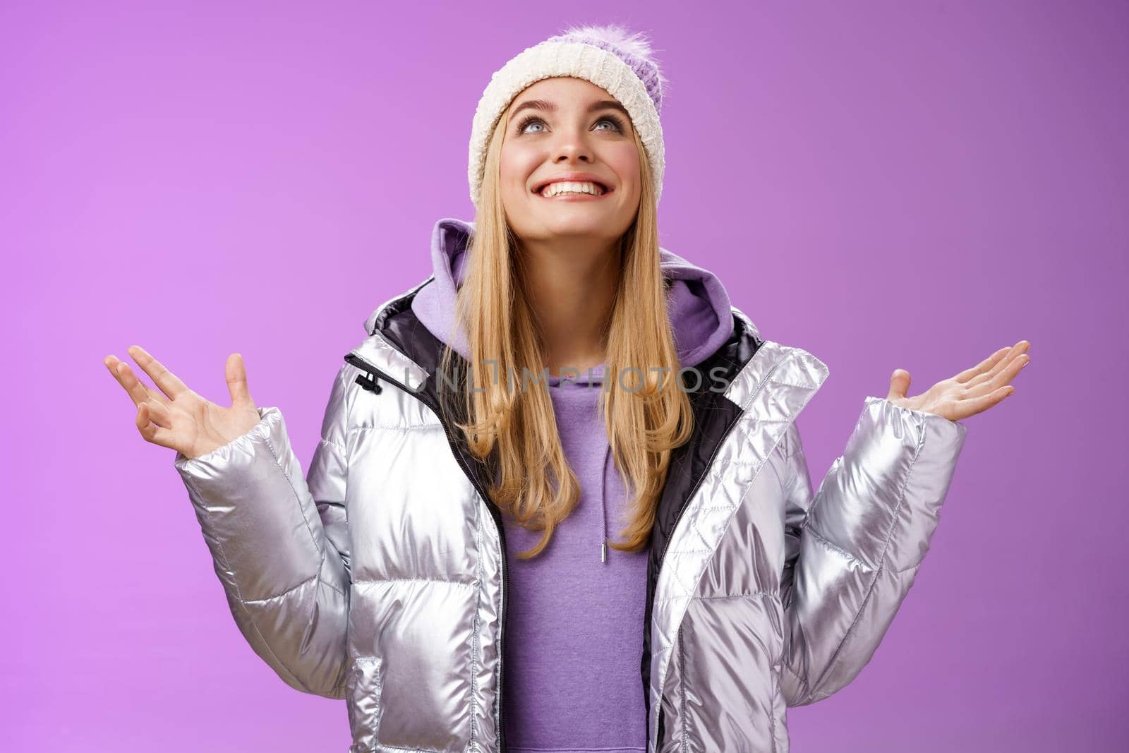 Thankful happy cute attractive blond young 25s woman in winter hat silver trendy jacket raise hands look up grateful god dream come true smiling delighted fulfilled wish, purple background.