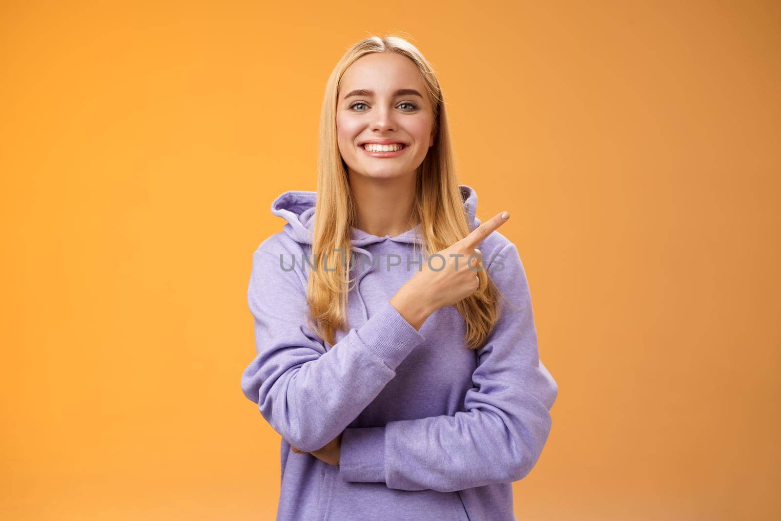 Charismatic modern millennial cute blond woman in hoodie smiling happily discussing cool new promo pointing right recommend check out visit interesting store near corner, orange background.