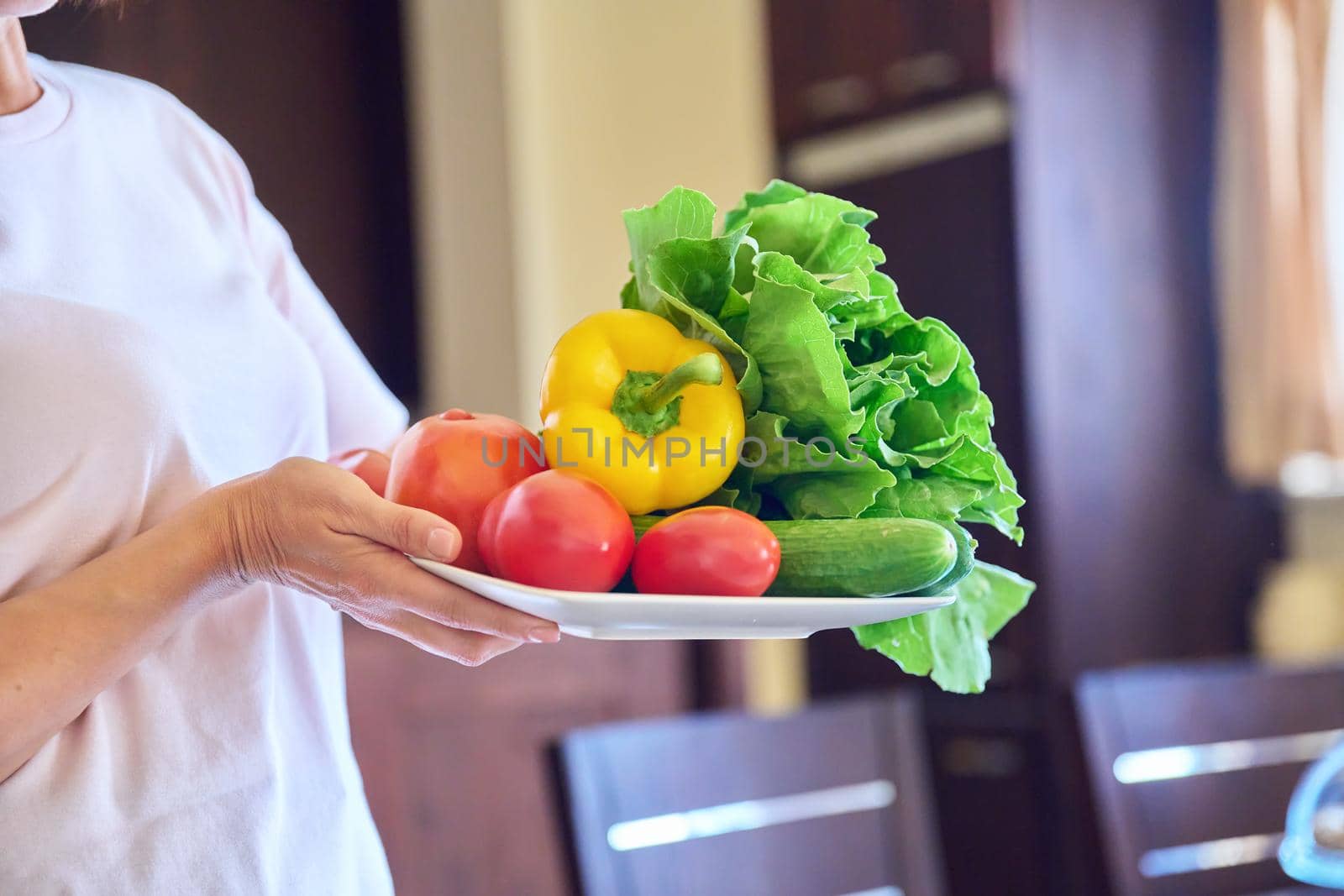 Close-up of a plate with tomatoes, cucumbers, paprika and lettuce in the hands of a woman. Kitchen interior background, fresh organic vegetables, home cooking, vegetarianism, nutrition, diet concept