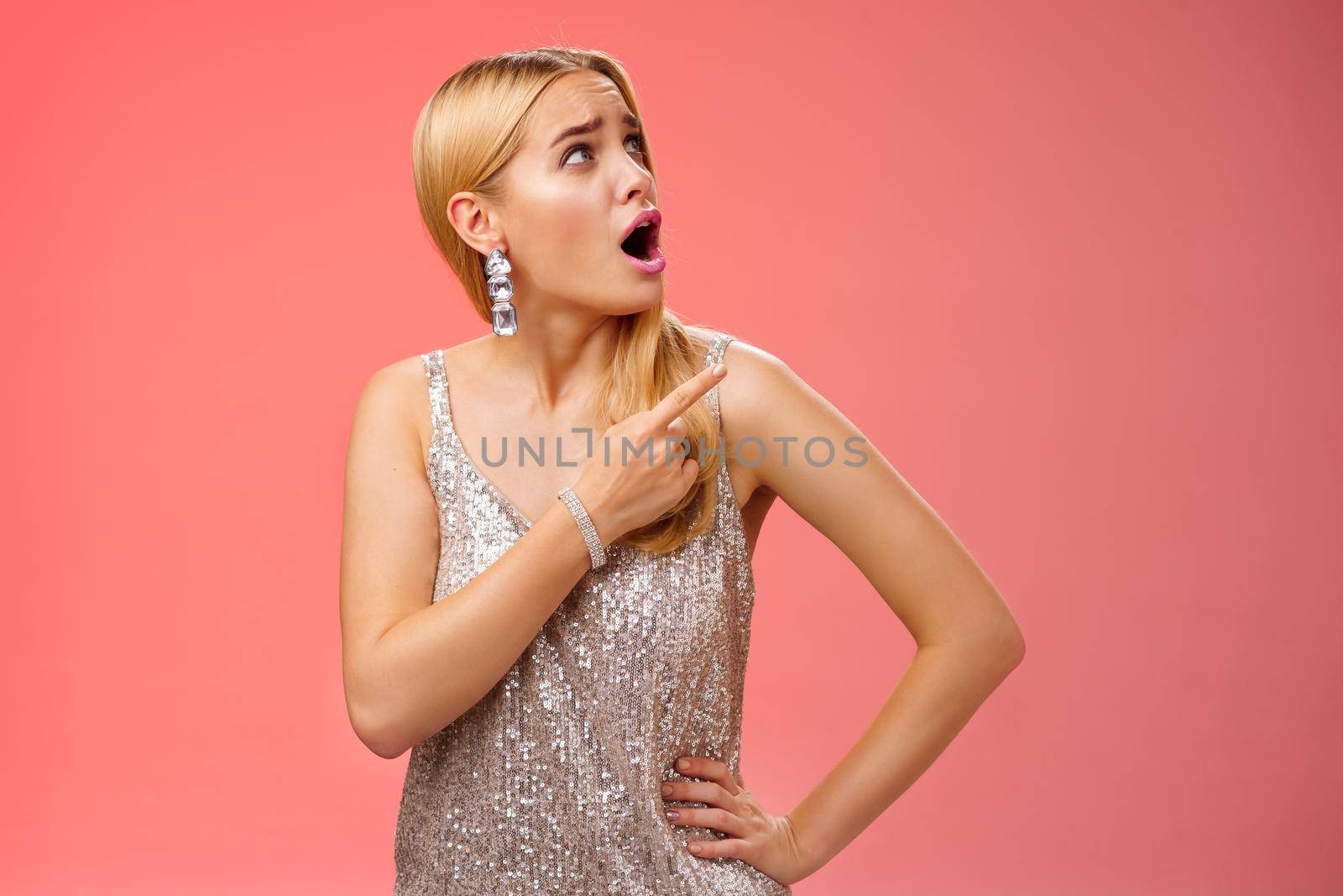 Shocked displeased bothered arrogant blond glamour woman in silver glittering dress turning upper right corner pointing complaining strange noise come upstairs, standing questioned red background.