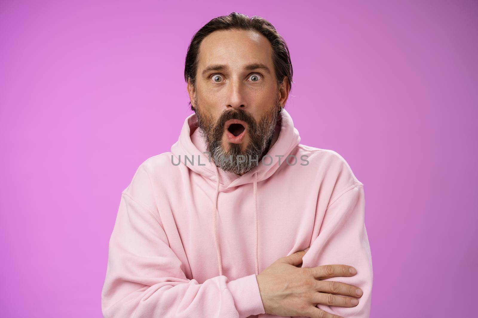 Shocked impressed gossiping caucasian bearded adult 40s man grey hair in pink hoodie gasping fascinated fold lips wow widen eyes astonished hearing interesting thrilling story, purple background.