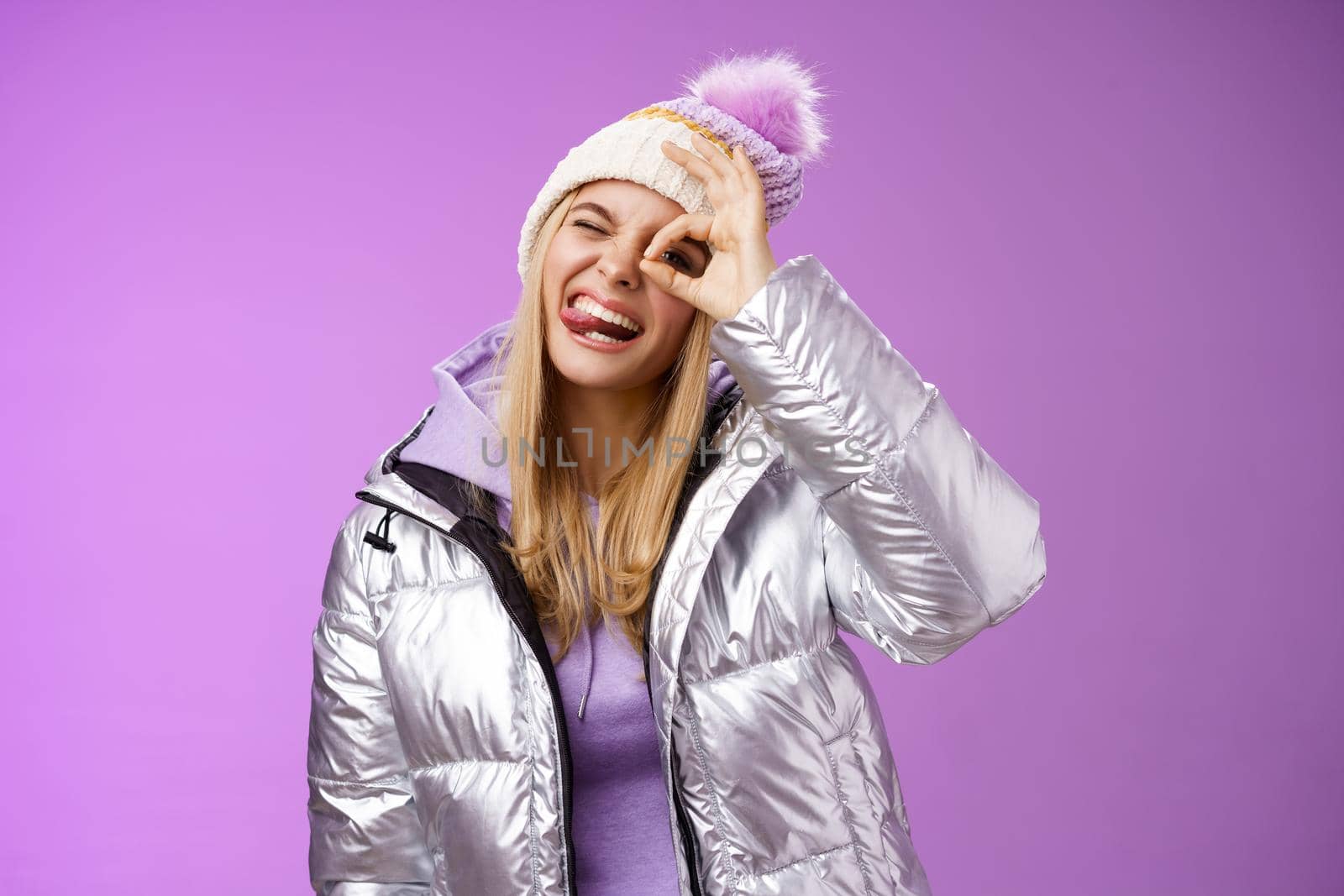 Friendly joyful caucasian blond girl in hat warm stylish shiny silver jacket tilt head happily show tongue smiling broadly enjoying awesome ski resort vacation travelling winter, purple background.