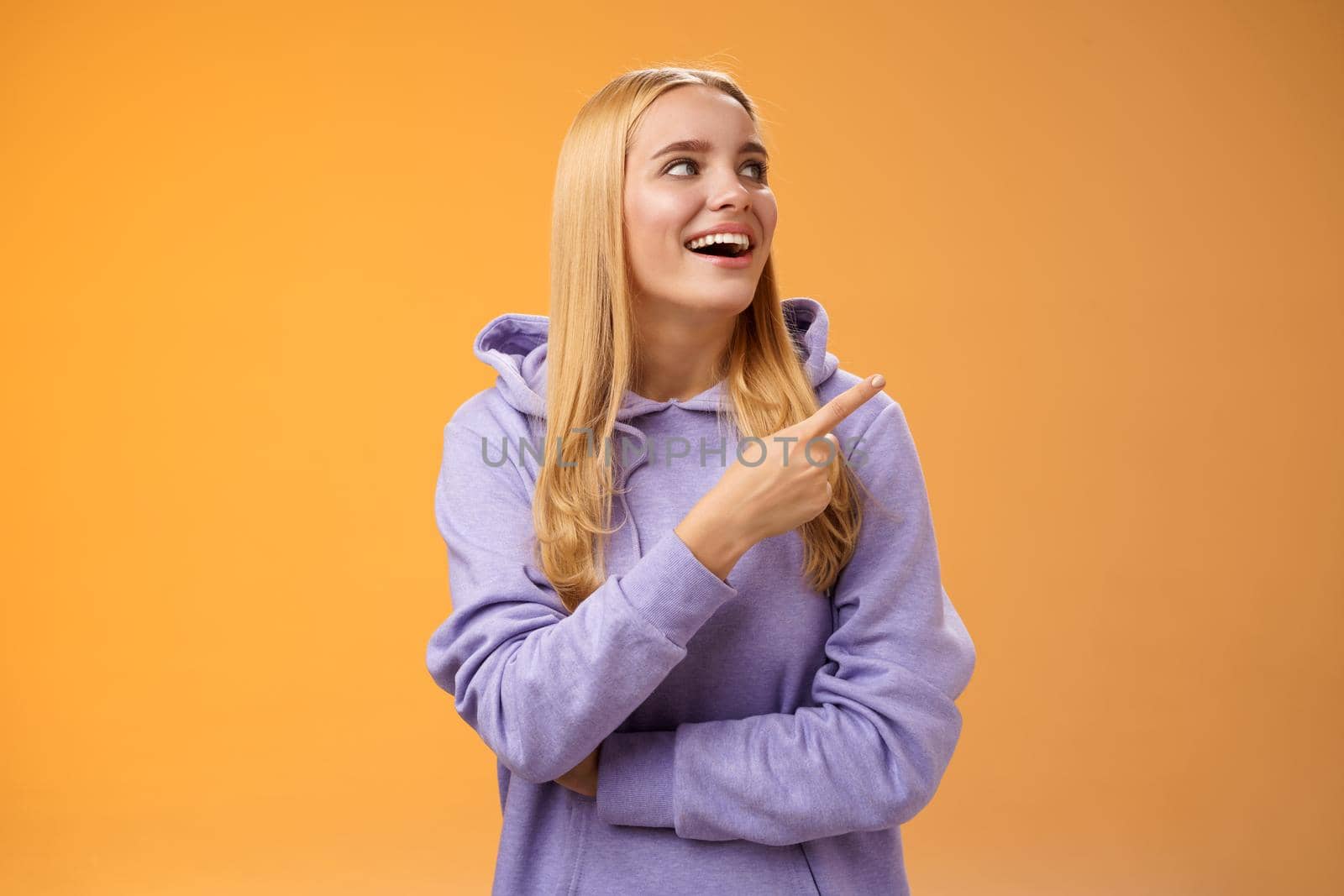 Amused carefree joyful blond european woman in hoodie pointing looking right pleased smiling happily enjoying interesting fascinating peromance making choice in store shopping, orange background.