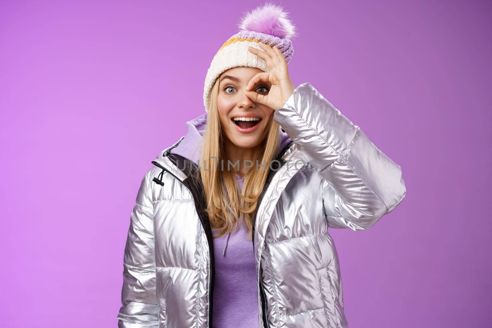 Impressed curious good-looking blond stylish female traveler charmed snowy mountains view hold okay ok sign eye smiling astonished speechless observing beautiful scenery in shiny silver jacket hat by Benzoix