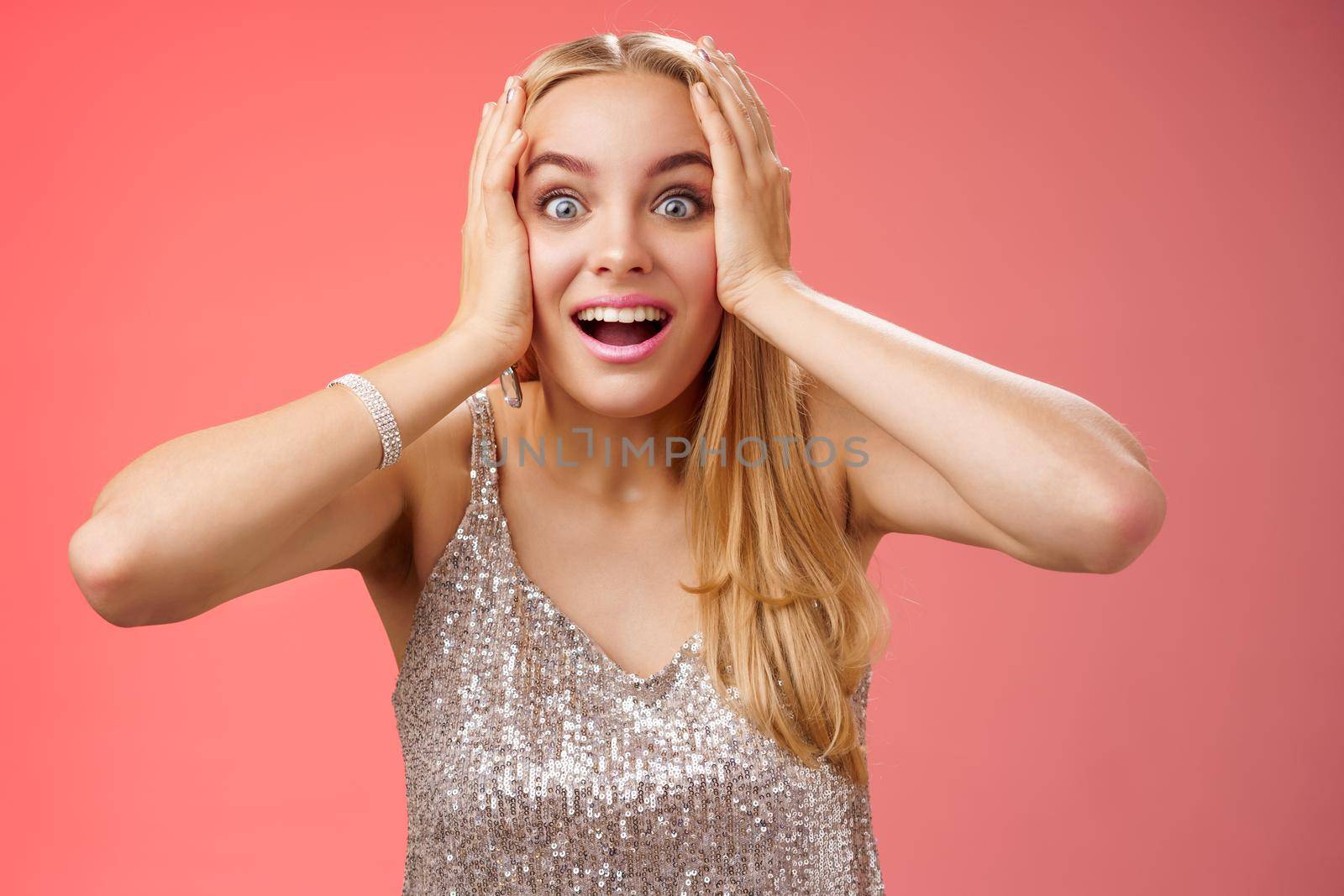 Amused surprised excited young blond attractive woman in silver glittering stylish dress hold head hands widen eyes speechless look camera unbelievable thrill fan see famous person, red background.