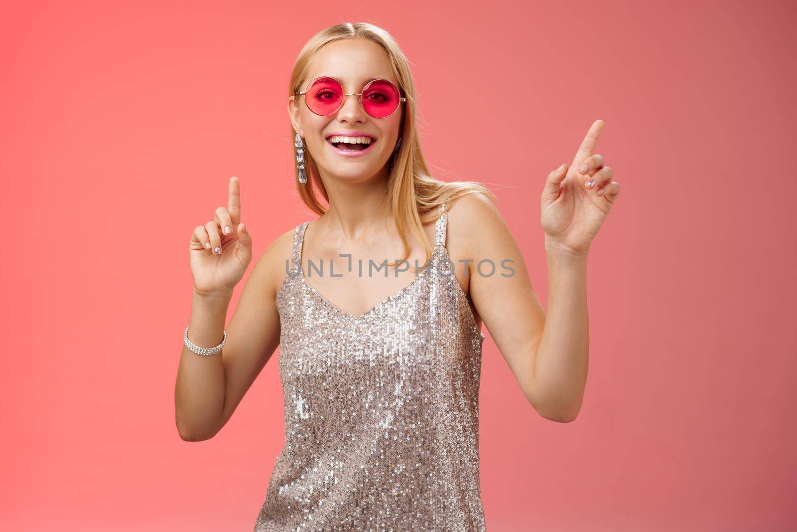 Stylish carefree fabulous young blond woman in silver dress red sunglasses having fun dancing joyfully dance-floor nightclub smiling broadly enjoying spend time friends, standing studio background.