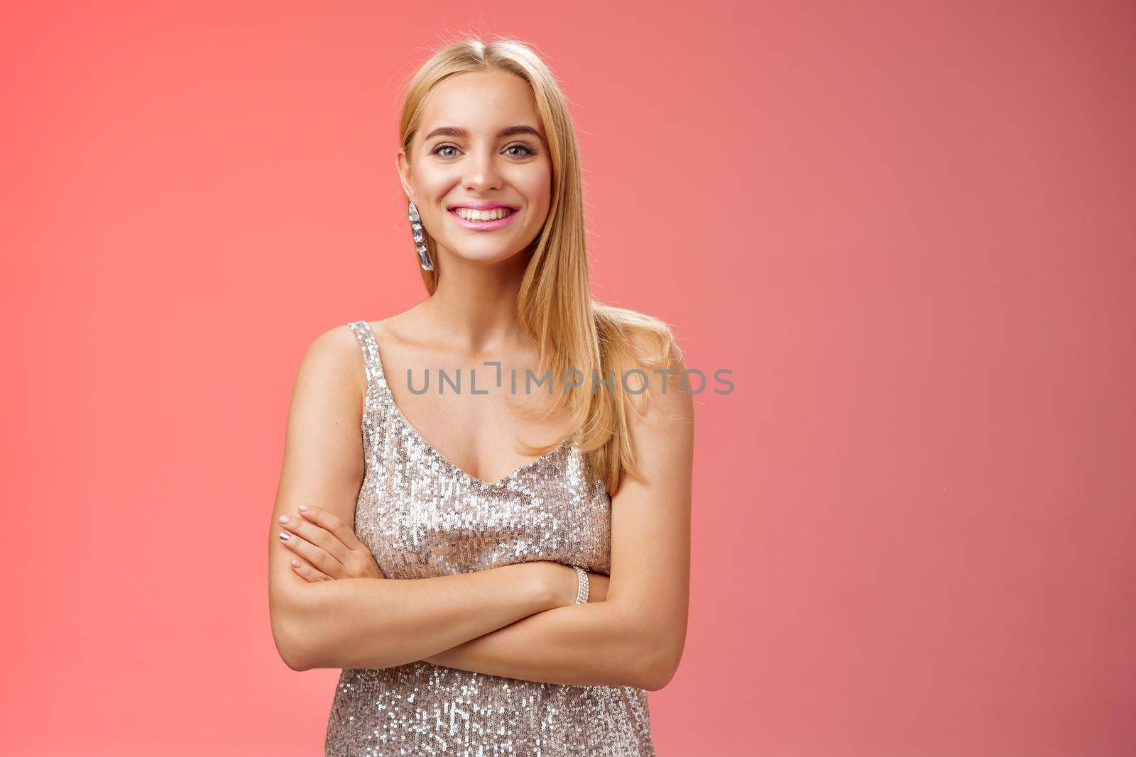 Charming tender blond girlfriend in silver shiny elegant dress feel little cold standing prom night talking boyfriend dance floor hold hand crossed chest grinning having fun, standing red background.