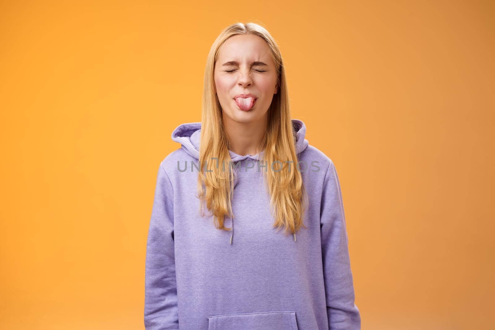 Playful amused charismatic young funny blond girl having fun close eyes showing tongue rebellious fool around unbothered enjoying carefree sunny spring days standing orange background.