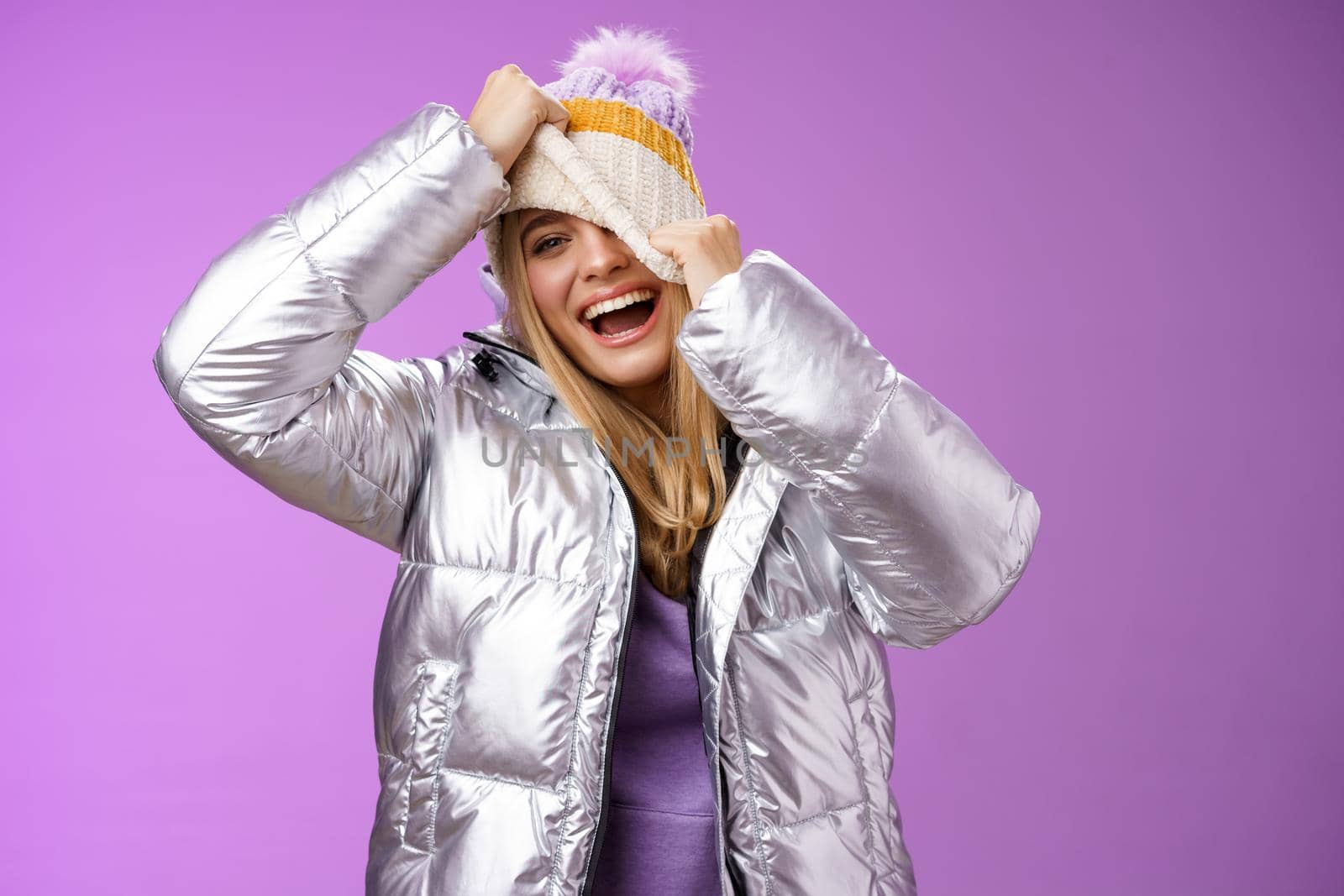 Amused carefree happy smiling playful attractive woman having fun laughing joyfully pulling hat face hiding peeking one eye camera grinning chuckling fool around have fun, purple background by Benzoix