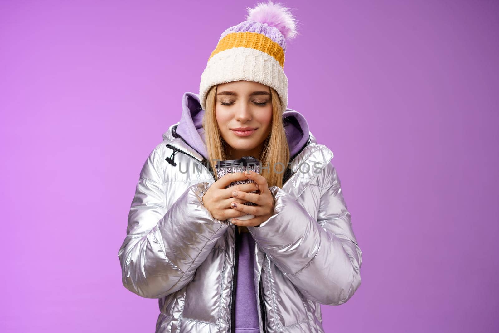 Charming tender romantic blond girl sniffing nice smell warm tasty coffee holding take-away cup warm hands close eyes look delighted wearing winter jacket hat enjoy vacation, purple background by Benzoix