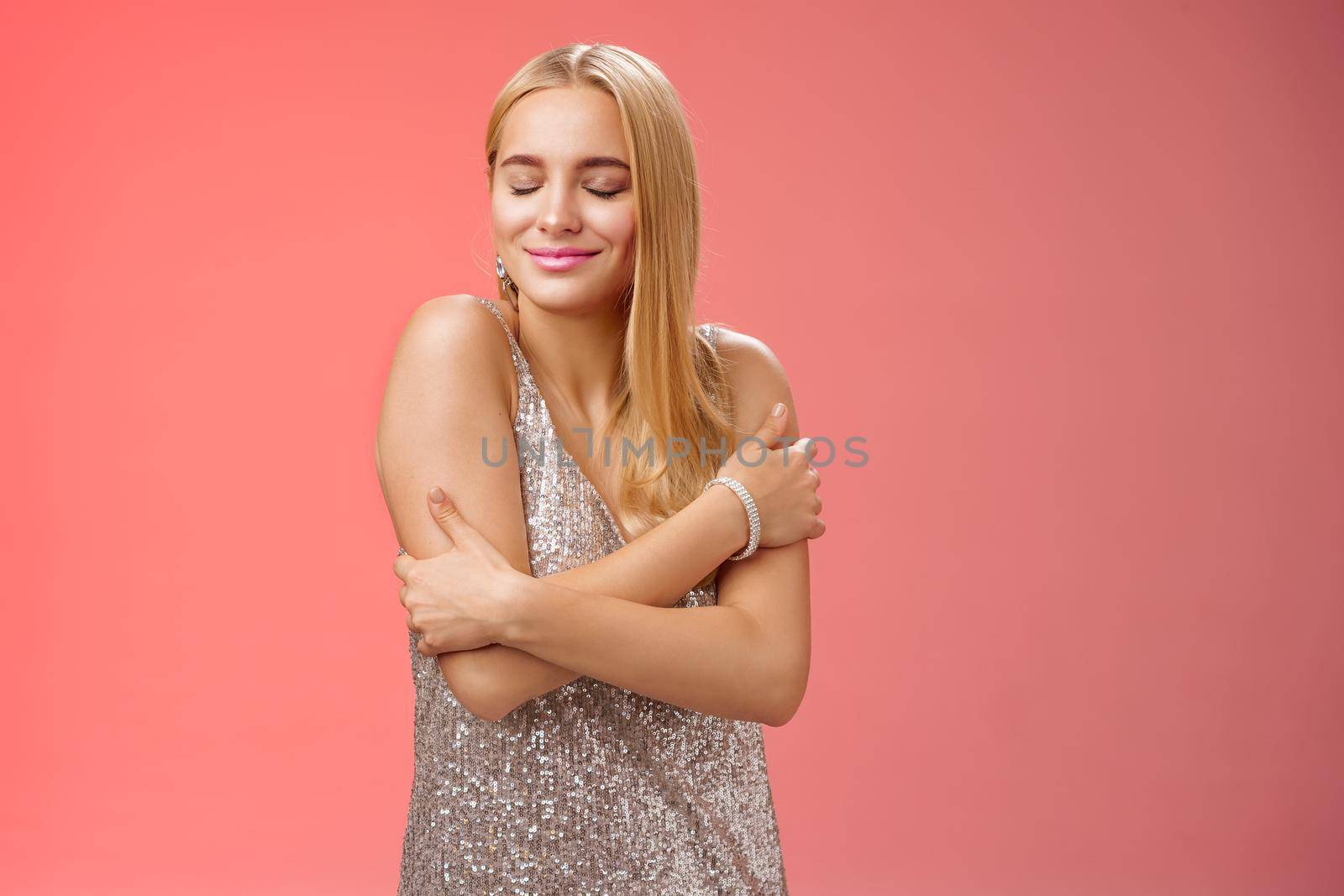 Lifestyle. Tender feminine elegant blond young woman in stylish silver glittering dress close eyes dreamy smiling touching herself cuddle embrace hands crossed body remember heartwarming passionate feelings.