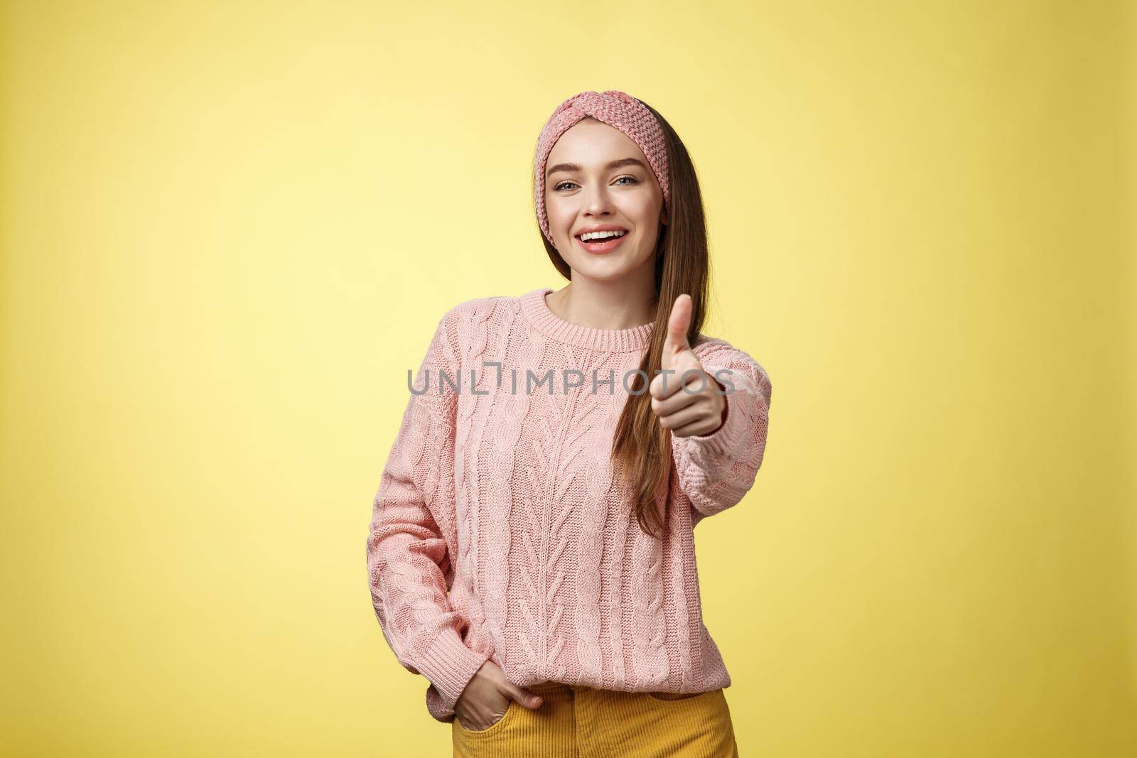Gestures, emotions and lifestlyle concept. Self-assured positive 20s european woman wearing casual sweater over yellow background showing thumb up gesture, approving, accepting and liking concept.