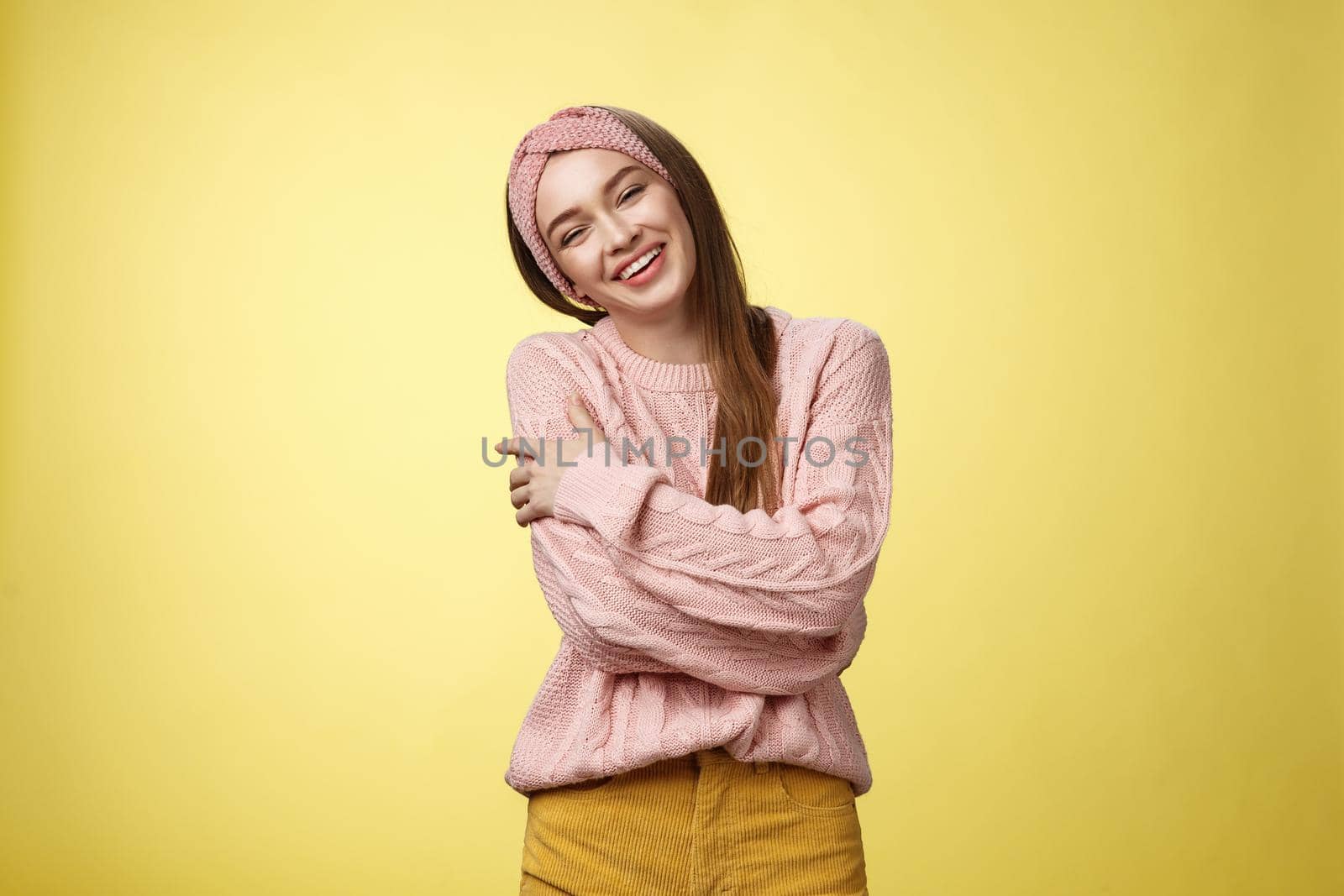 Cozy charming friendly young smiling girl in knitted warm comfortable sweater, grinning joyfully tilt head emracing herself, crossing arms, hugging feeling safe and happy, posing against yellow wall.