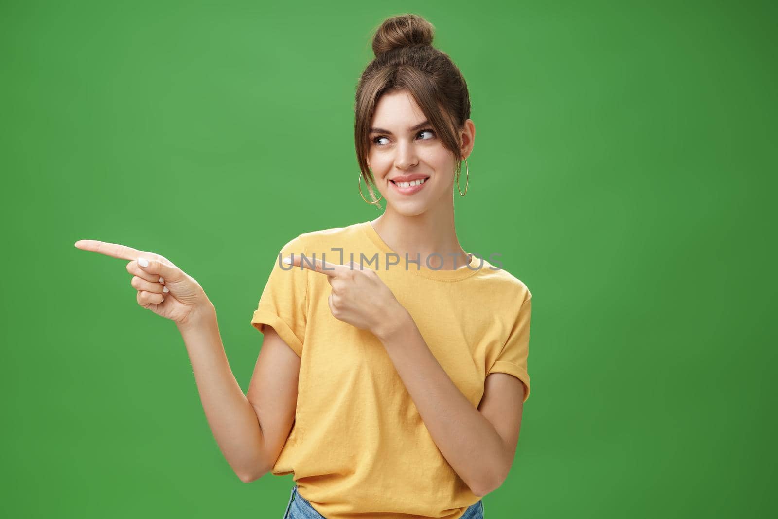 Waist-up shot of charismatic happy and carefree charming woman in yellow t-shirt pointing and looking left enthusiastic and pleased smiling cheerfully posing against green background delighted. Copy space