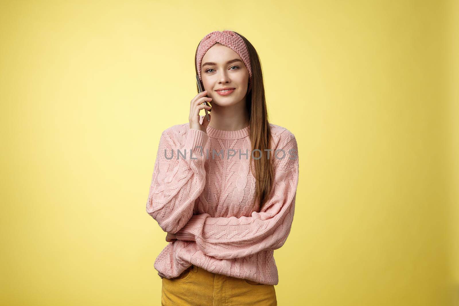 Stylish young girlfriend having pleasant conversation via phone holding smartphone near ear talking, making reservation in restaurant, ordering food delivery, smiling, posing against yellow wall by Benzoix