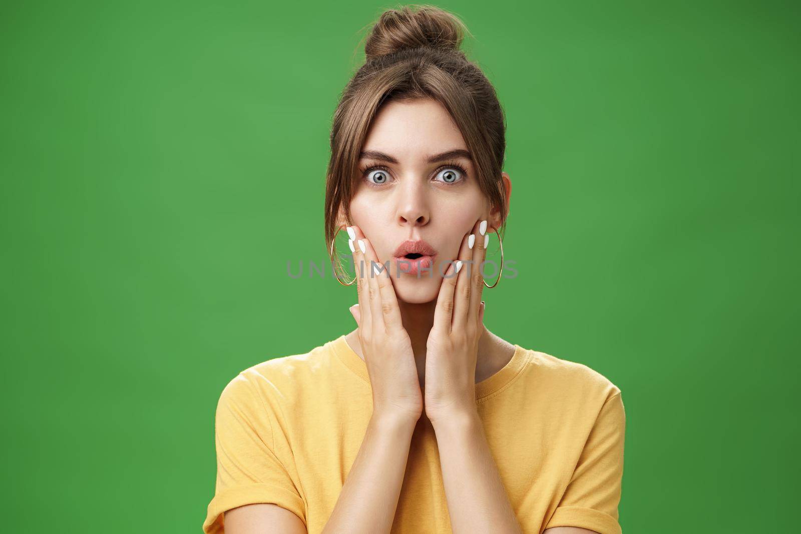 Portrait of impressed and shocked cute feminine woman in yellow t-shirt folding lips from excitement and interest touching cheeks surprised reacting to amazing rumor posing over green background. Body language concept