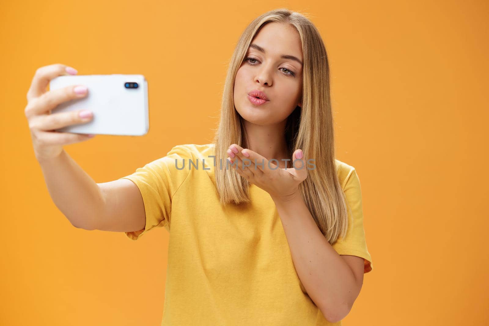 Stylish glamourous female fashion blogger ending recording video via smartphone by sending air kiss at camera, taking selfie with sensual and confident gaze at screen posing over orange wall. Lifestyle, blogger, van life concept