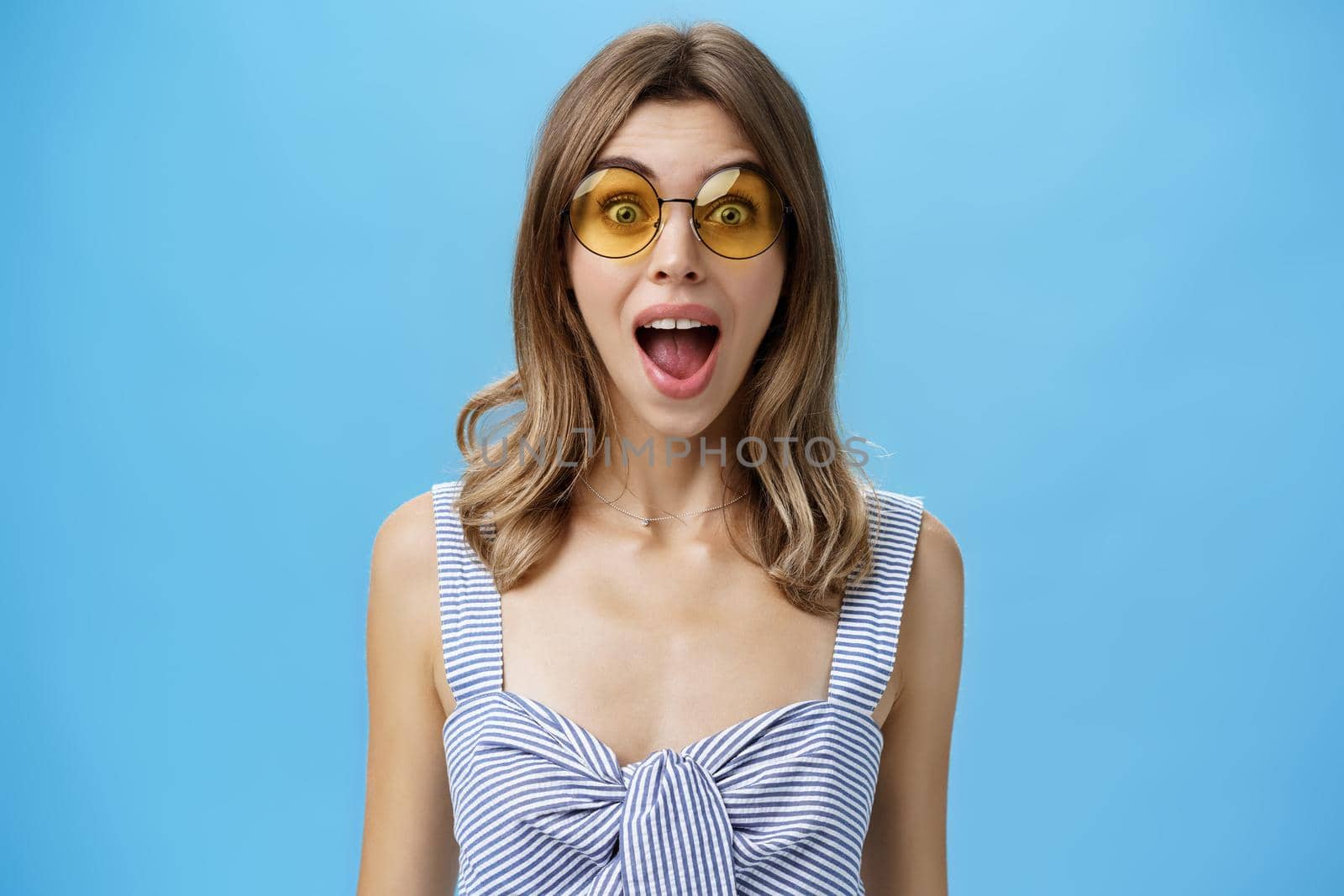 Waist-up shot of excited surprised and emotive charismatic caucasian woman in trendy sunglasses opening mouth from amazement and joy gazing at camera impressed and astonished over blue background. Lifestyle.