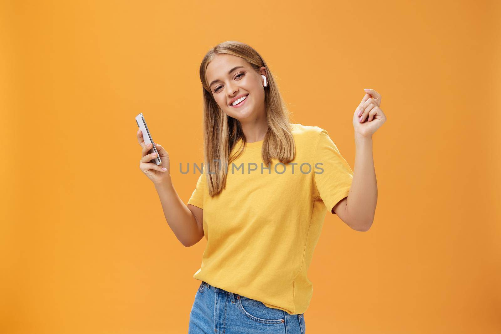 Joyful carefree european woman wearing wireless earbuds holding smartphone dancing with raised hands and pleased happy smile leading vanlife having time to relax and enjoy life over orange wall. Technology concept