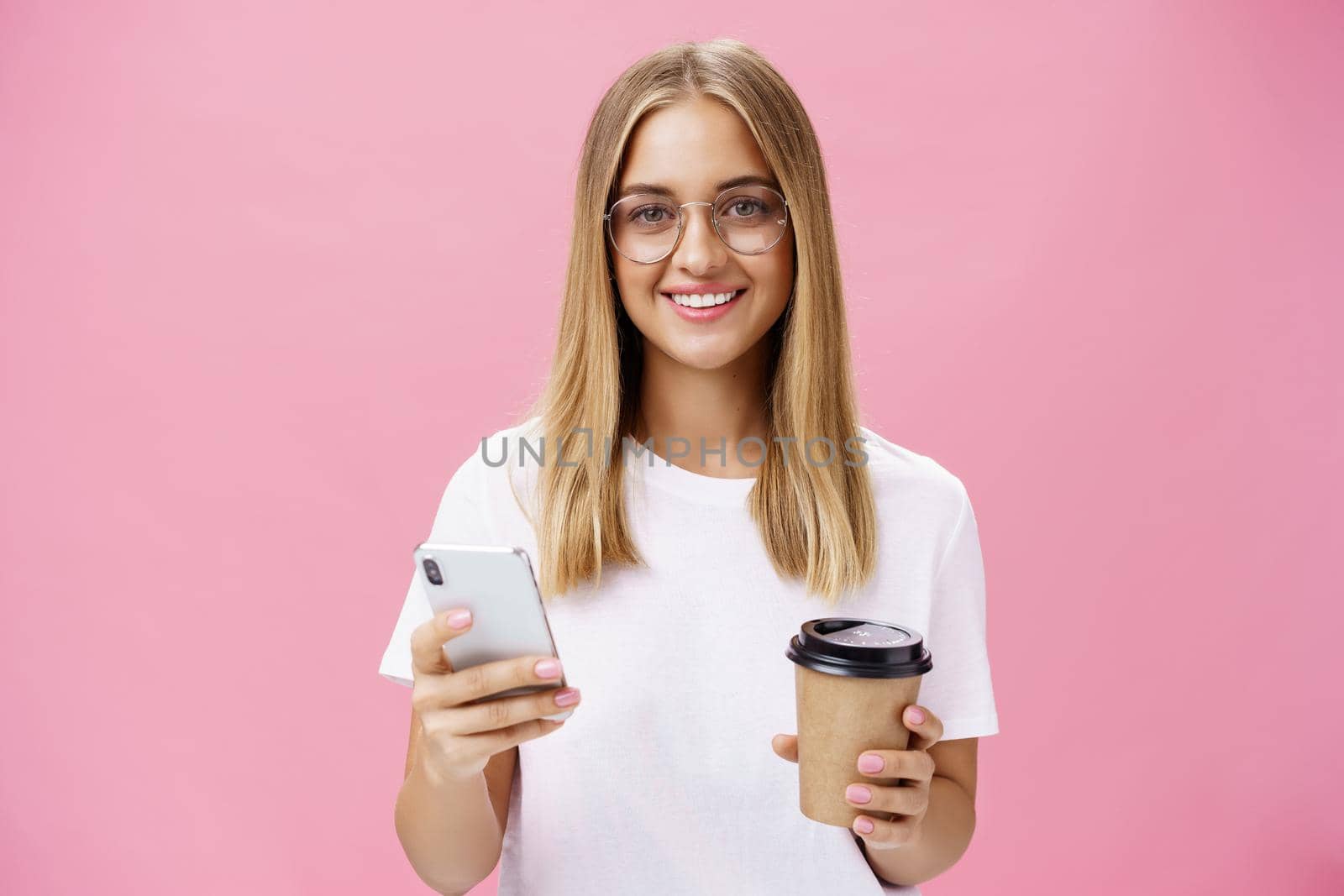 Pleasant friendly-looking girl smiling at camera holding paper cup of coffee and smartphone. Portrait of joyful nice woman drinking morning drink, posing opinion about cafe in internet over pink wall. Technology and lifestyle concept