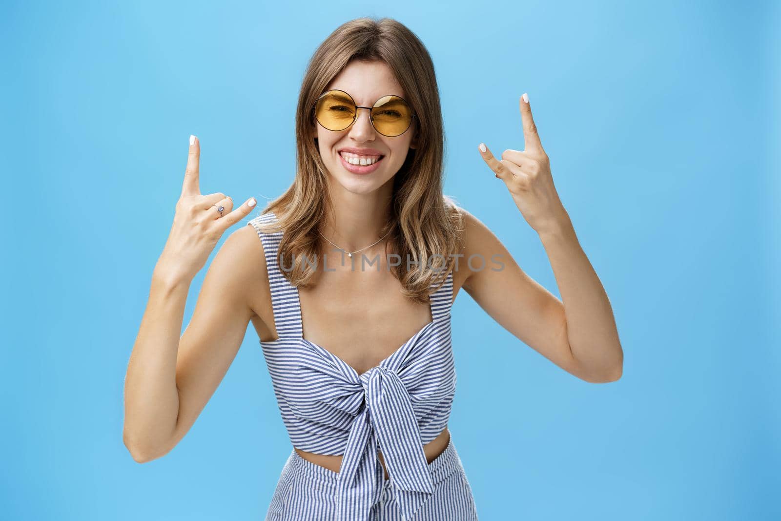 Girl ready to rock-n-roll this party wearing best matching outfit from wardrobe and cool sunglasses showing rock gestures, smiling excited having fun listening music on concert over blue wall.