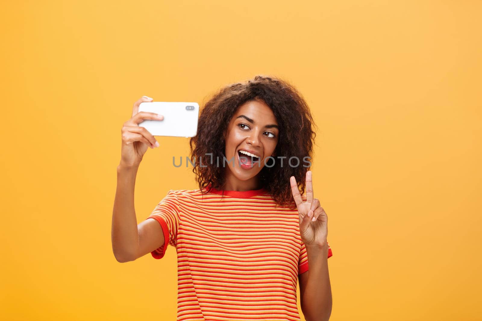 Smile to camera. Attractive and stylish self-assured dark-skinned female model with curly hairstyle showing peace gesture while taking selfie holding smartphone near face and smiling at gadget screen by Benzoix