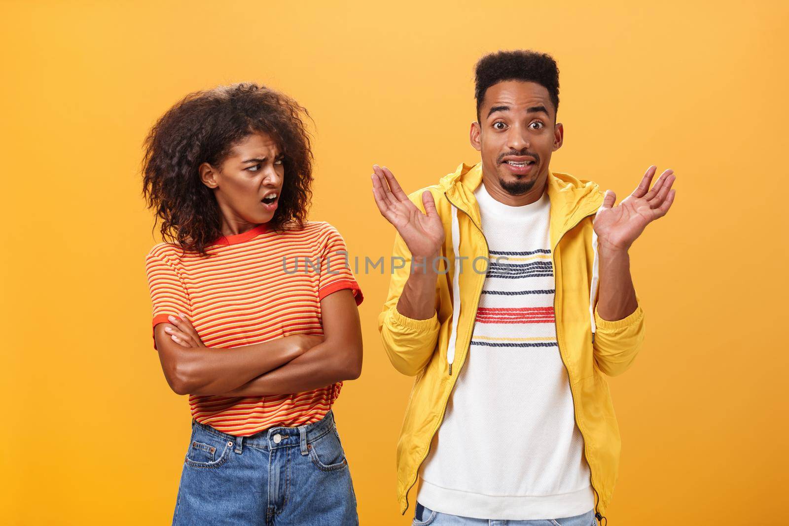 Jeez man what your problem. Offended displeased african american stylish and attractive girlfriend looking at boyfriend acting cool pretending he uninvolved shrugging with sorry look and smiling by Benzoix