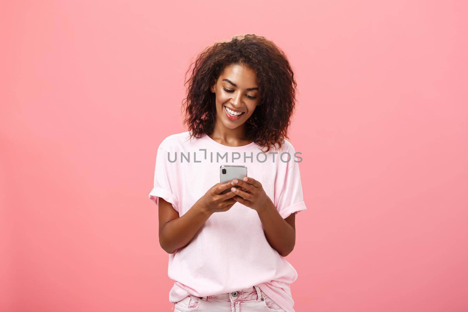 Girl writing text message to friend. Portrait of charming amused and happy young dark-skinned woman with curly hair looking at smartphone screen typing smiling enjoying cool features of cellphone by Benzoix