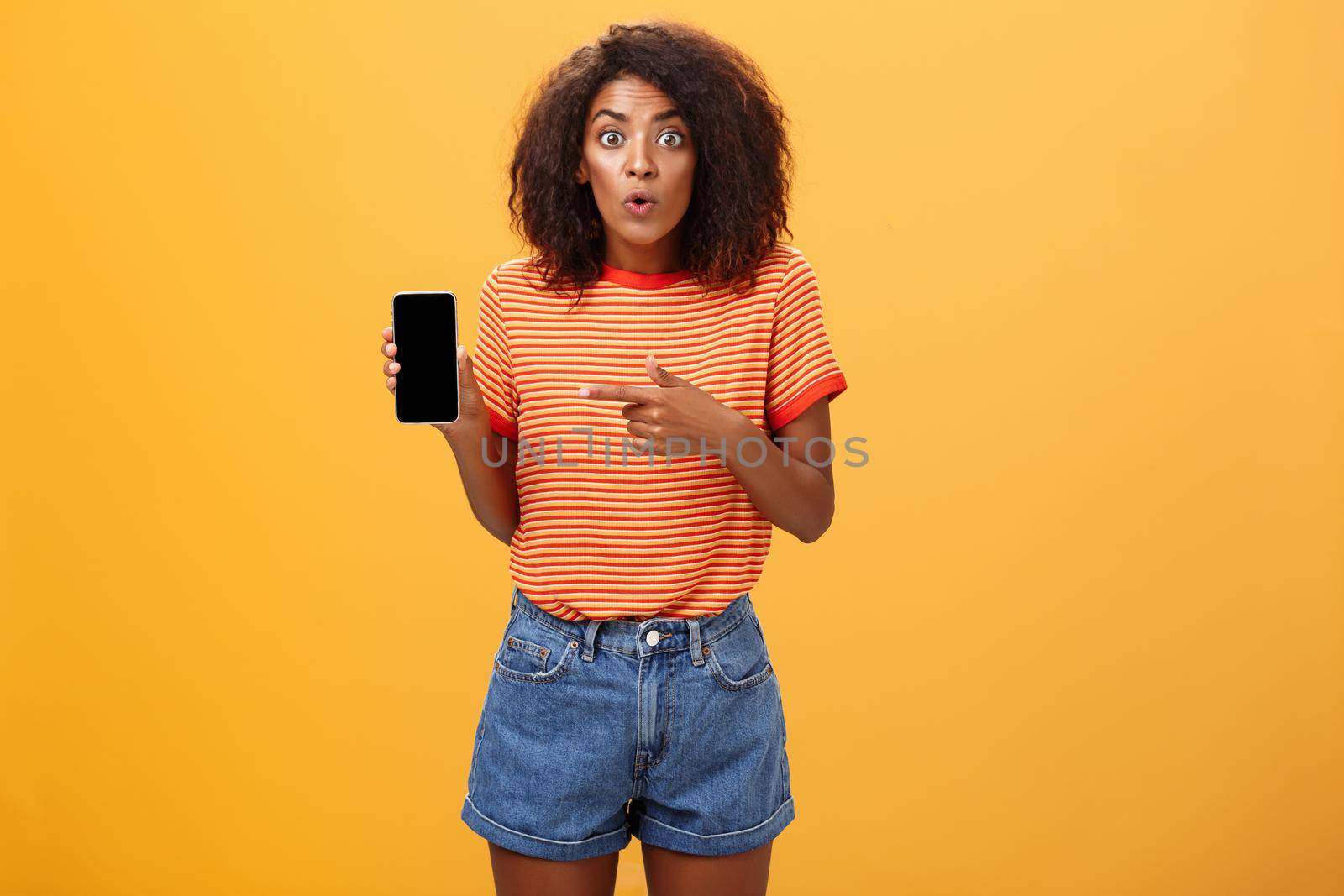 Portrait of impressed and curious female friend showing profile of ex to friend talking about shocking changes of appearance folding lips from interest holding smartphone pointing at cellphone screen by Benzoix