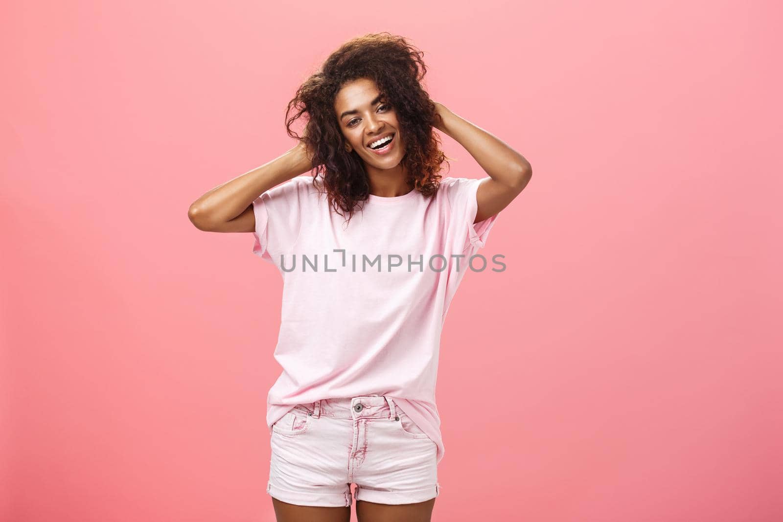 Girl enjoying nice weather chilling making photos for social network touching curly hair gently and carefree smiling broadly at camera standing over pink background happy and chill. Emotions and people concept