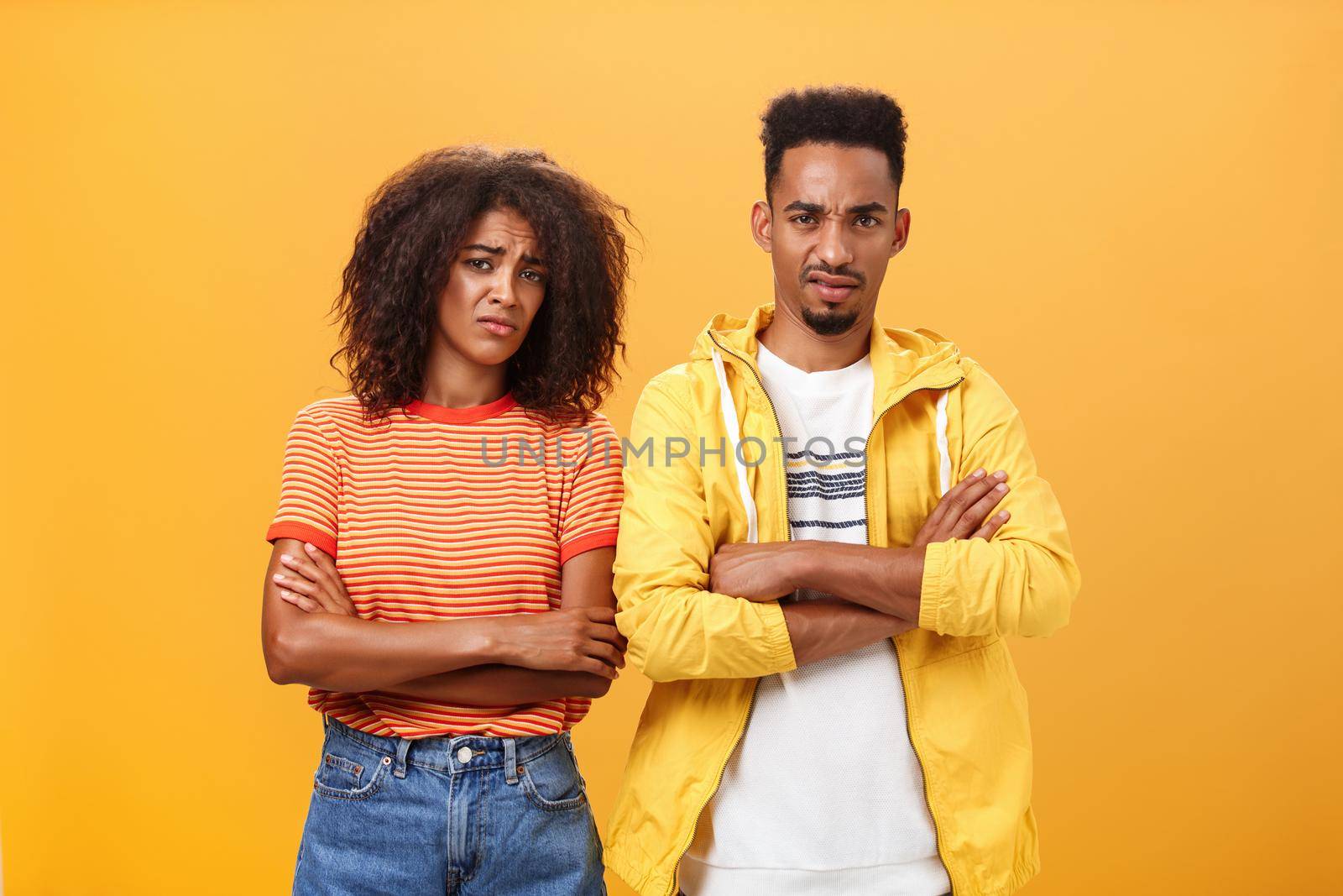 Two friends dislike lame idea of mate. Portrait of dissatisfied unimpressed african american man and woman crossing arms on chest in aversion frowning doubtful and disappointed over orange wall by Benzoix