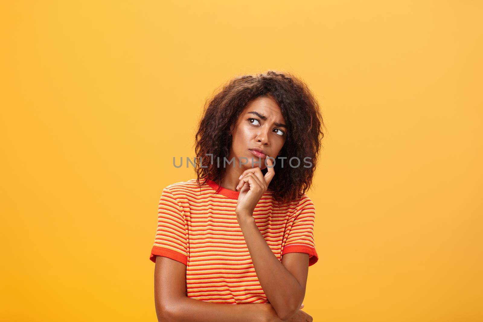Woman considering to take part in event. Smart focused and creative good-looking african girl with curly hair holding finger on lip gazing concerned left, thinking making decision over orange wall.
