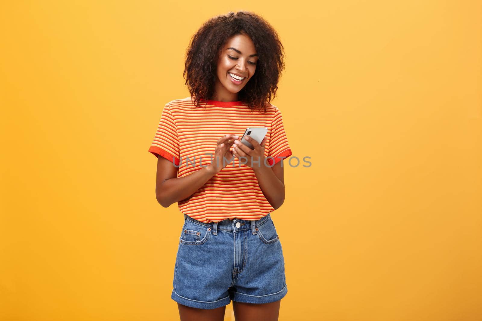 Stylish carefree girl texting friend come over standing pleased over orange wall in stylish denim shorts typing message or scrolling news in internet via smartphone gazing at device screen with smile by Benzoix