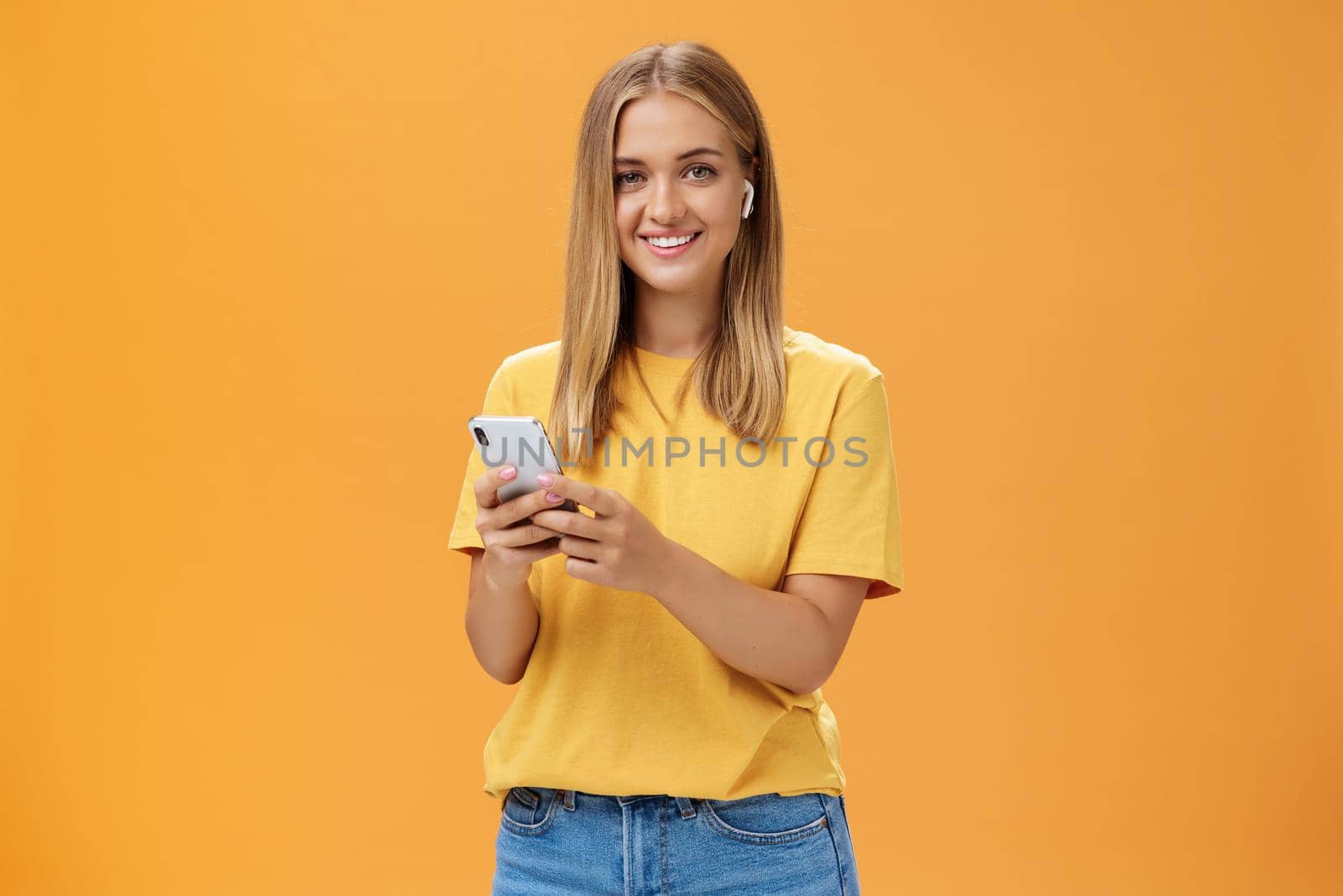 Young caucasian girl with tanned skin and fair hair using wireless earphones to call friend via smartphone holding cellphone against chest, smiling cheerfully at camera getting used to new technology by Benzoix
