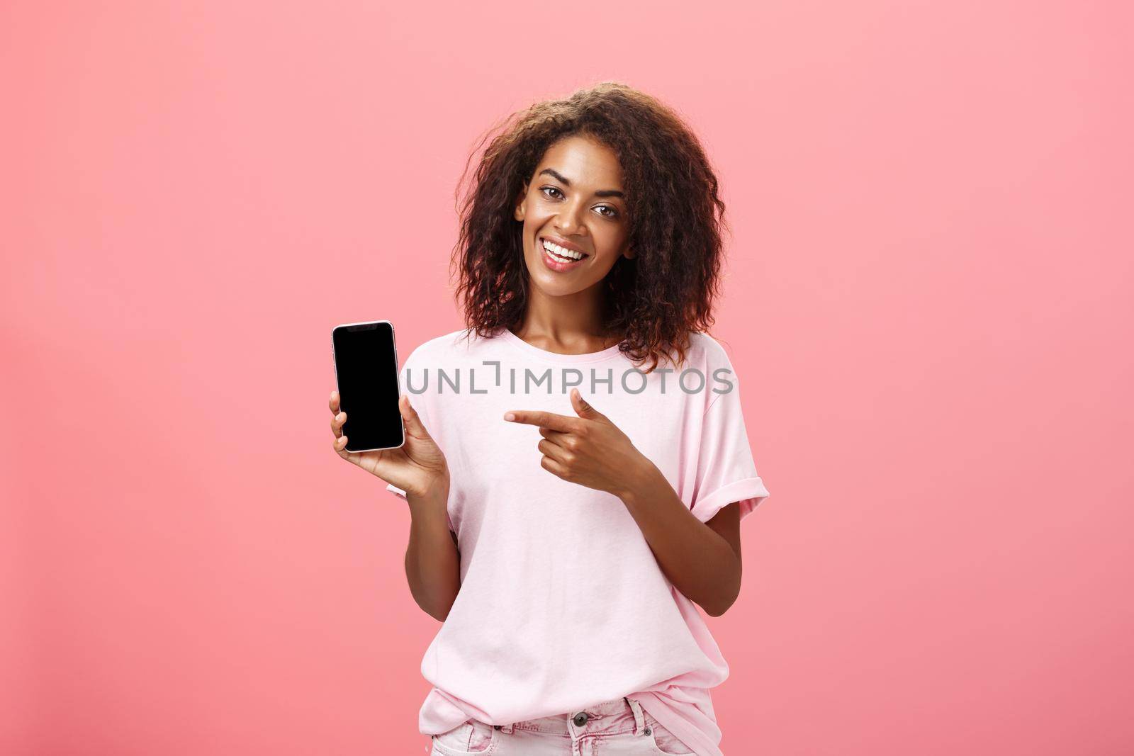 Suggest this model of phone. Happy charming african american curly-haired woman in stylish outfit holding smarpthone and pointing at device screen while recommending cool app over pink wall.