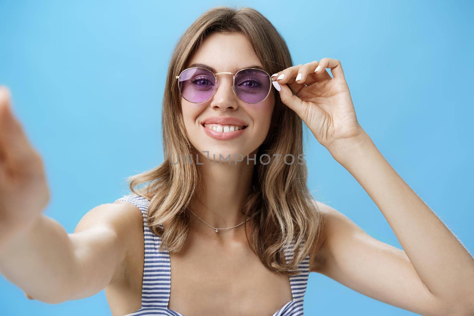Charming carefree european female in trendy sunglasses touching frames holding camera with one hand to take selfie smiling broadly feeling happy enjoying life on weekends over blue wall. Lifestyle.