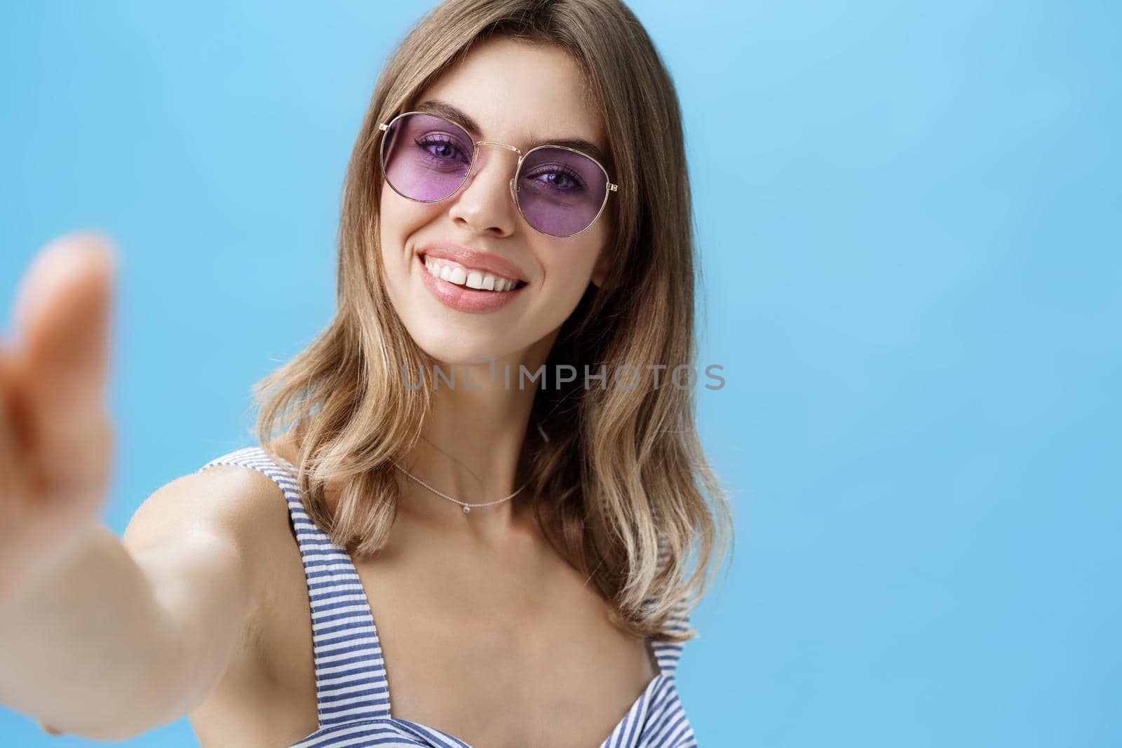 Charming woman with cute gapped teeth in sunglasses relaxing on vacation taking beautiful shots pulling hand towards camera holding it to take selfie smiling broadly over blue background by Benzoix