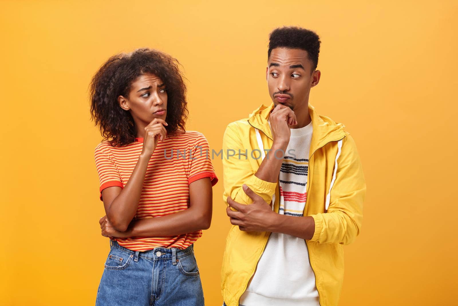 Silly confused girl standing gloomy and worried pursing lips and frowning holding hand on chin looking at boyfriend confused while guy glancing at girlfriend with curious expression over orange wall. Lifestyle.